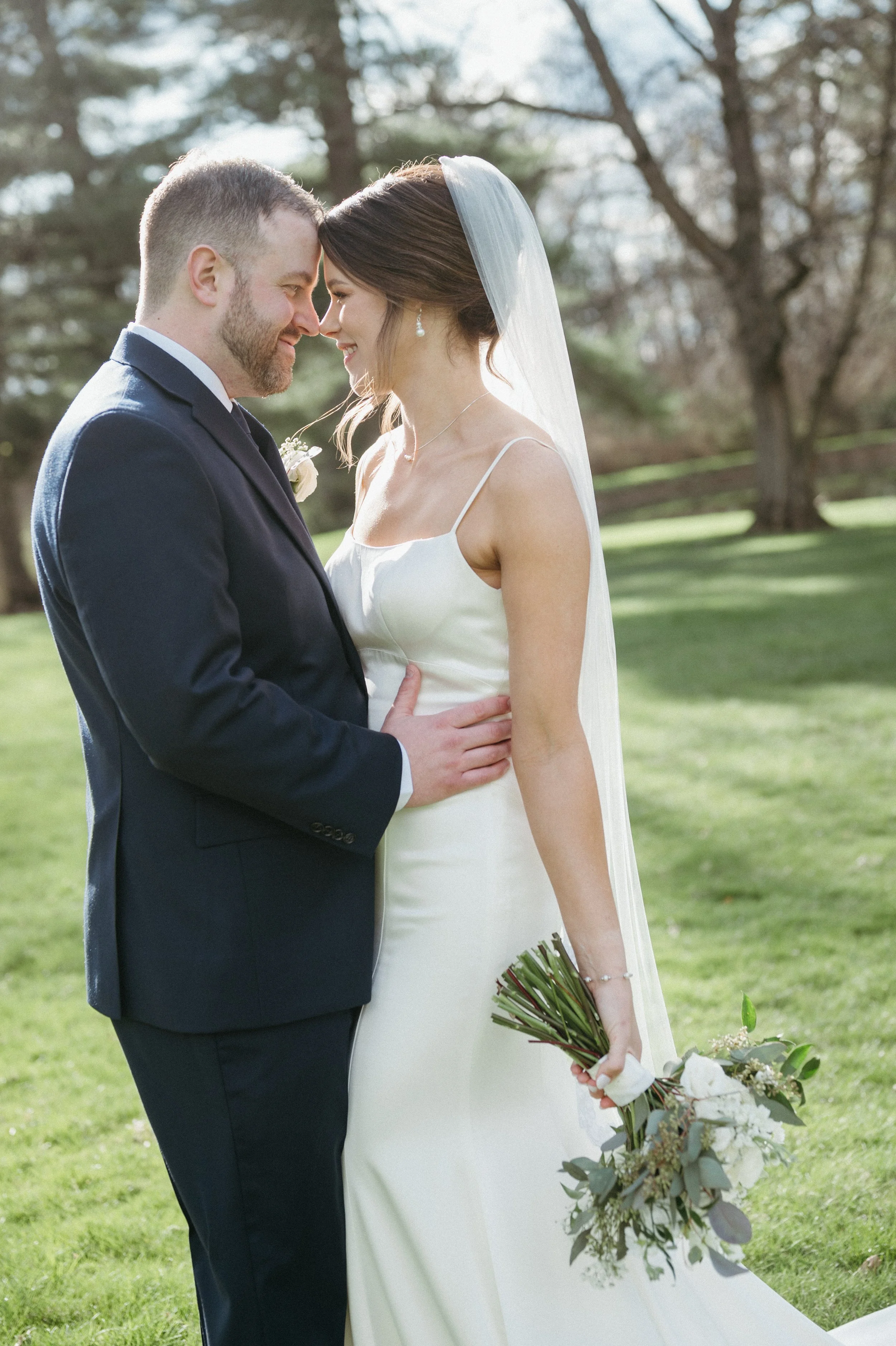 A bride and groom standing outdoors on a grassy area, embracing each other with big smiles, surrounded by green trees and foliage.