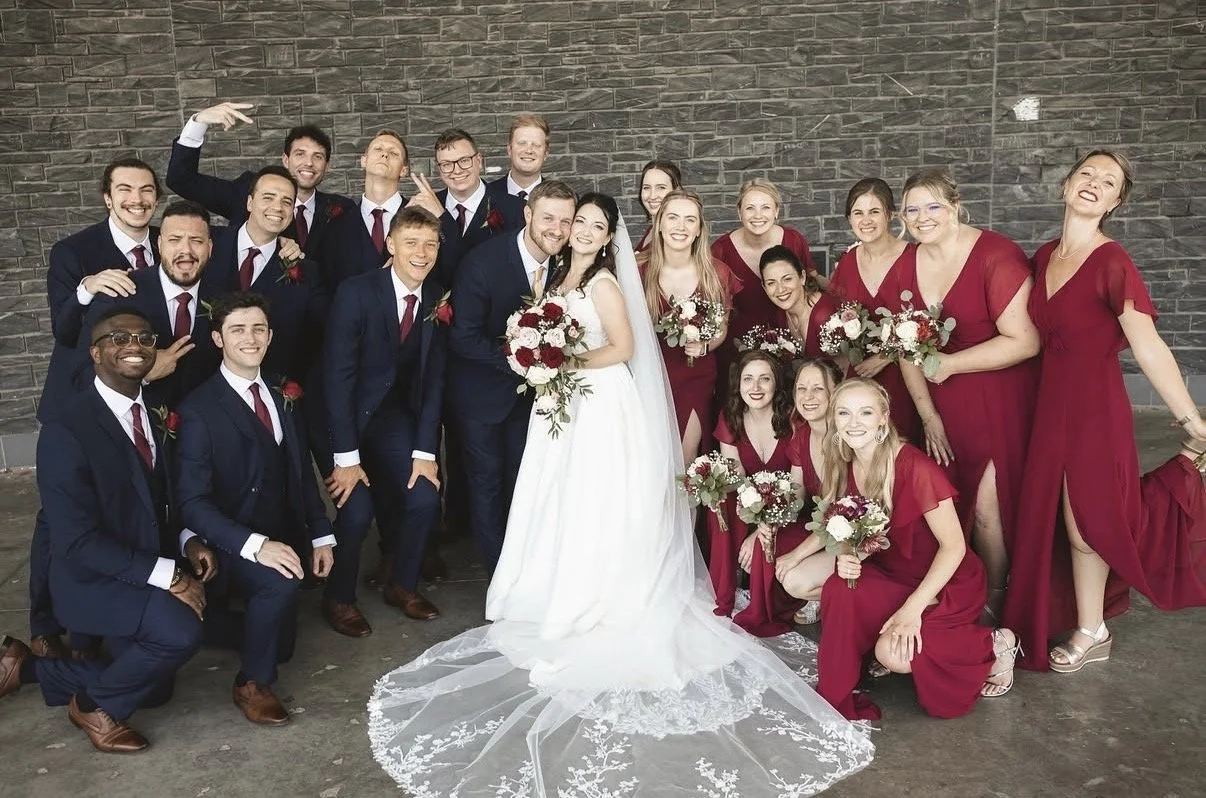 Group of wedding guests and bridal party posing outdoors against a brick wall, with a bride and groom in the center. The bride wears a white wedding dress with a long veil and holds a bouquet, while the groom wears a dark suit and boutonniere. Female guests wear red dresses, and male guests wear dark suits with red ties and boutonnieres. Everyone is smiling and celebrating.