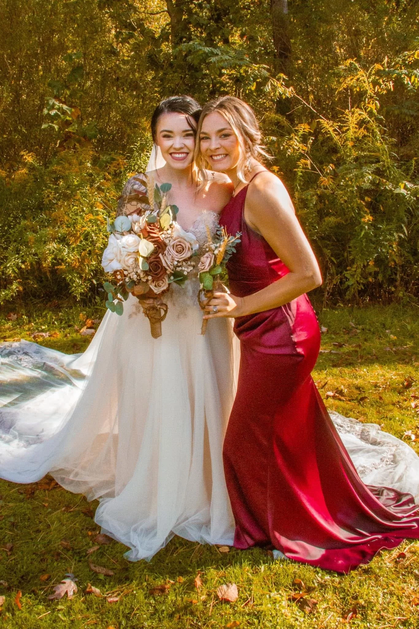 Two women, one in a wedding dress holding a bouquet, and the other in a burgundy evening gown, standing outdoors in a wooded area with sunlight filtering through the trees, smiling and posing for a photo.