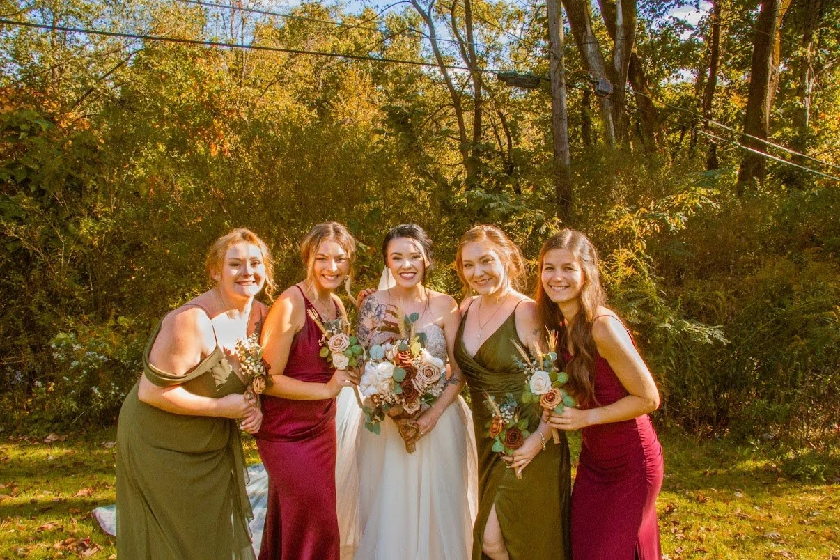 Five women in formal dresses standing outdoors, holding bouquets, during daytime. The woman in the center is wearing a white dress, likely a bride, with four women dressed in shades of green and burgundy surrounding her in a natural setting with trees and sunlight.