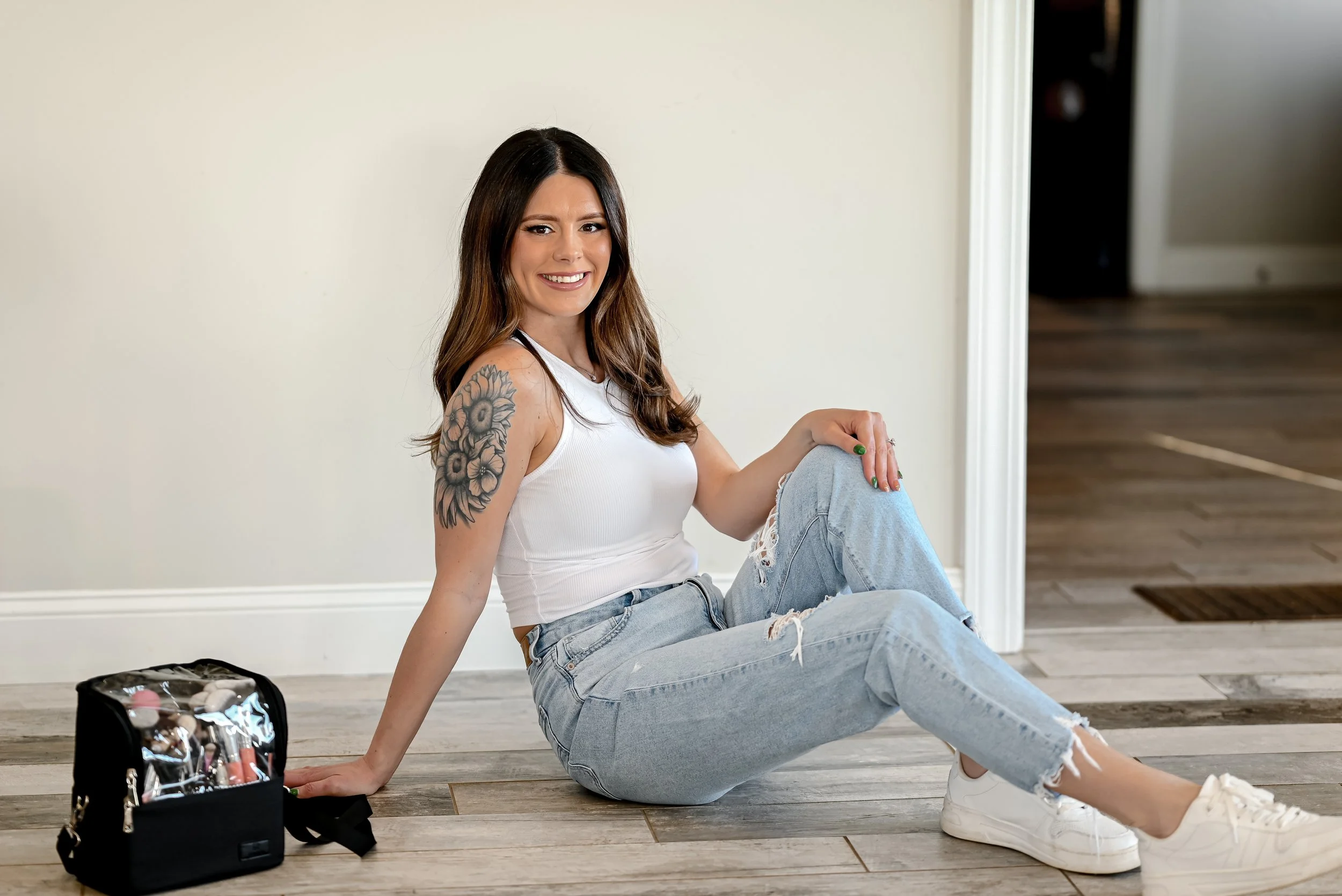 Young woman with long brown hair, white tank top, ripped jeans, and white sneakers, sitting on wooden floor next to a makeup bag, smiling at the camera.