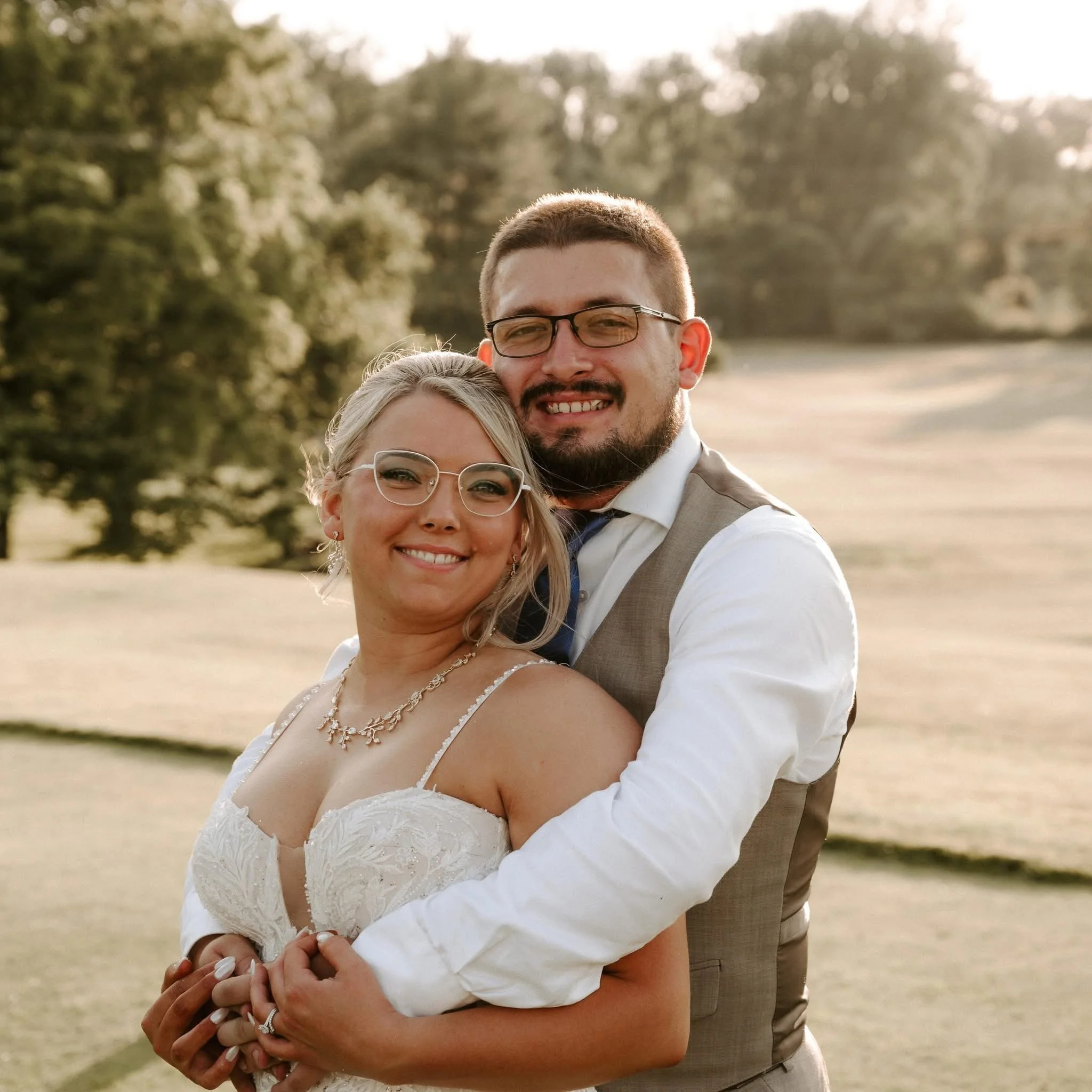 A smiling bride and groom standing outdoors on a grassy field during sunset, with trees in the background, dressed in wedding attire.