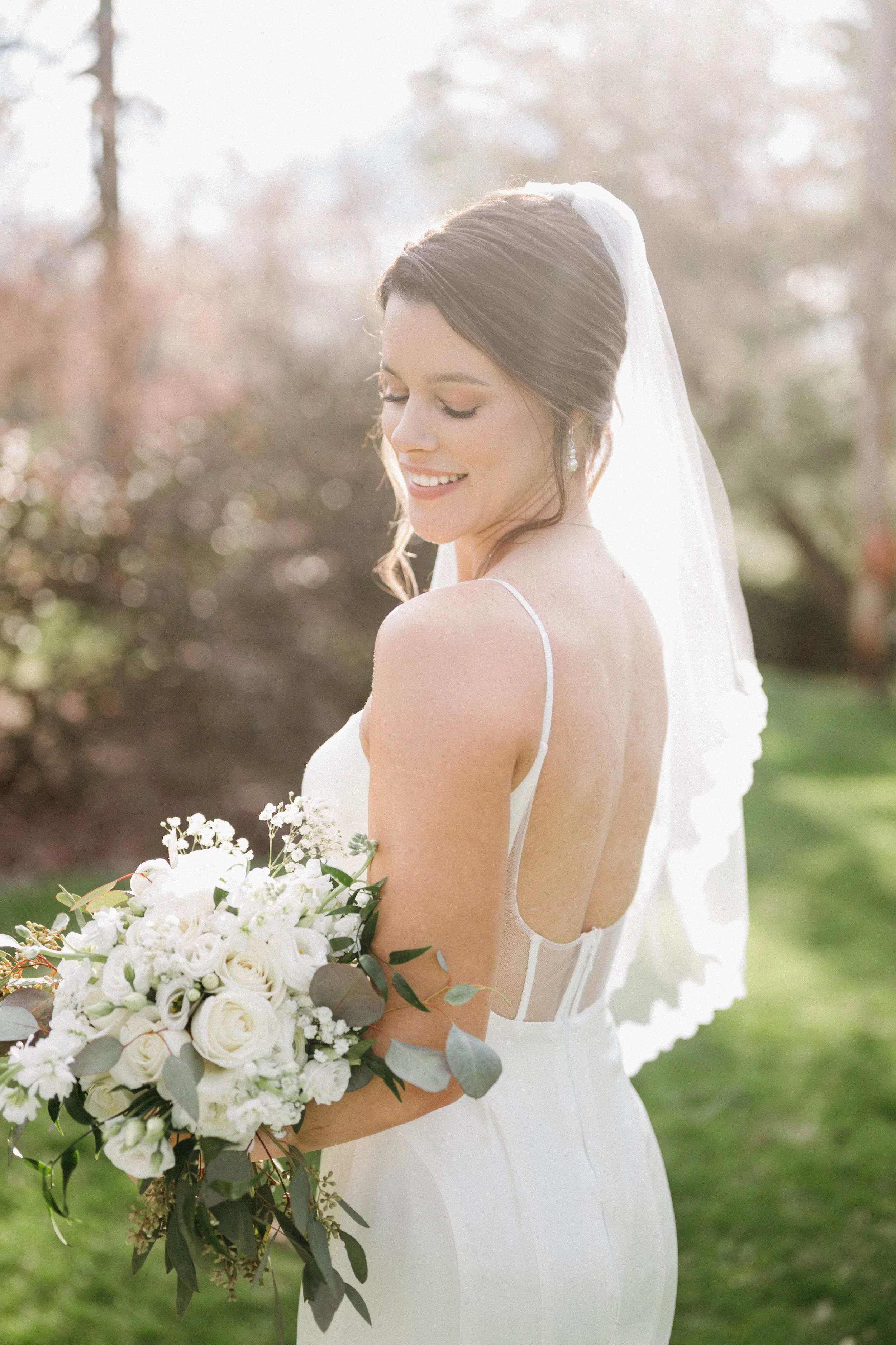 A bride holding a bouquet of white roses and greenery, smiling with her eyes closed outdoors.