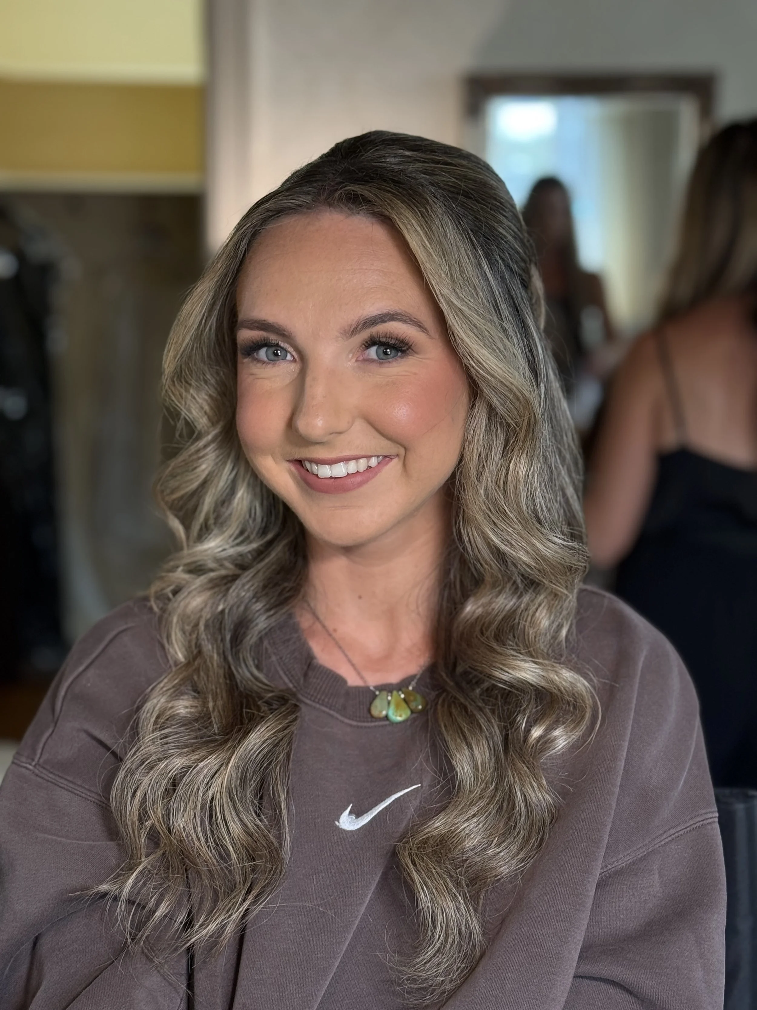 A young woman with long, wavy blonde hair and blue eyes smiling at the camera, wearing a brown Nike sweatshirt and a necklace, in a room with blurred background of other people.