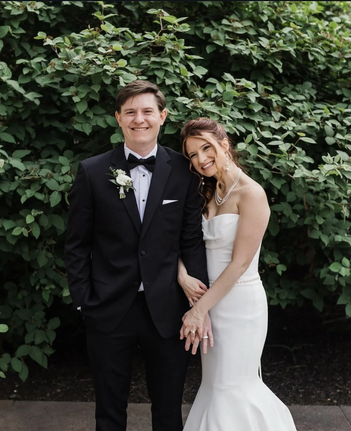 A smiling bride and groom standing outdoors in front of green foliage during their wedding. The groom is dressed in a black tuxedo with a bow tie and a white boutonniere. The bride is wearing a strapless white wedding gown with pearl jewelry, and has her hair styled with loose curls. They are holding hands and smiling happily.