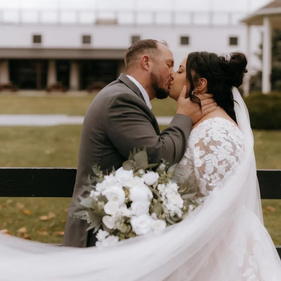 A bride and groom share a kiss outdoors on their wedding day, with the groom holding the bride's face and the bride holding a bouquet of white flowers.