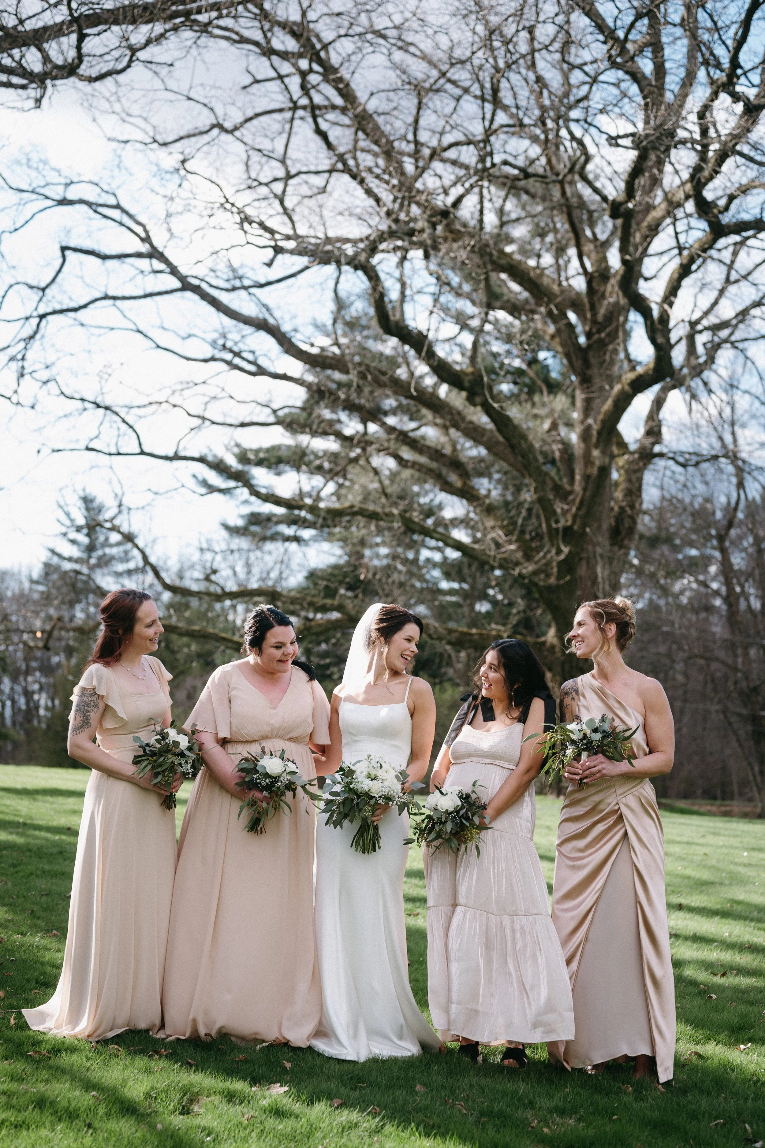 Five women in wedding dresses and bridesmaid dresses standing outdoors on green grass with a large tree behind them. They are smiling and holding bouquets of flowers.