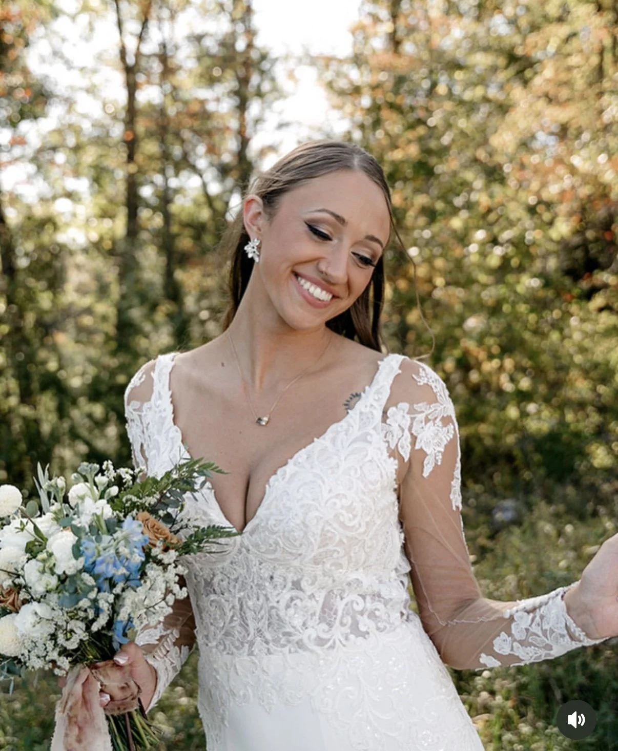 A smiling bride in a white lace wedding dress holding a bouquet of flowers outdoors during autumn.