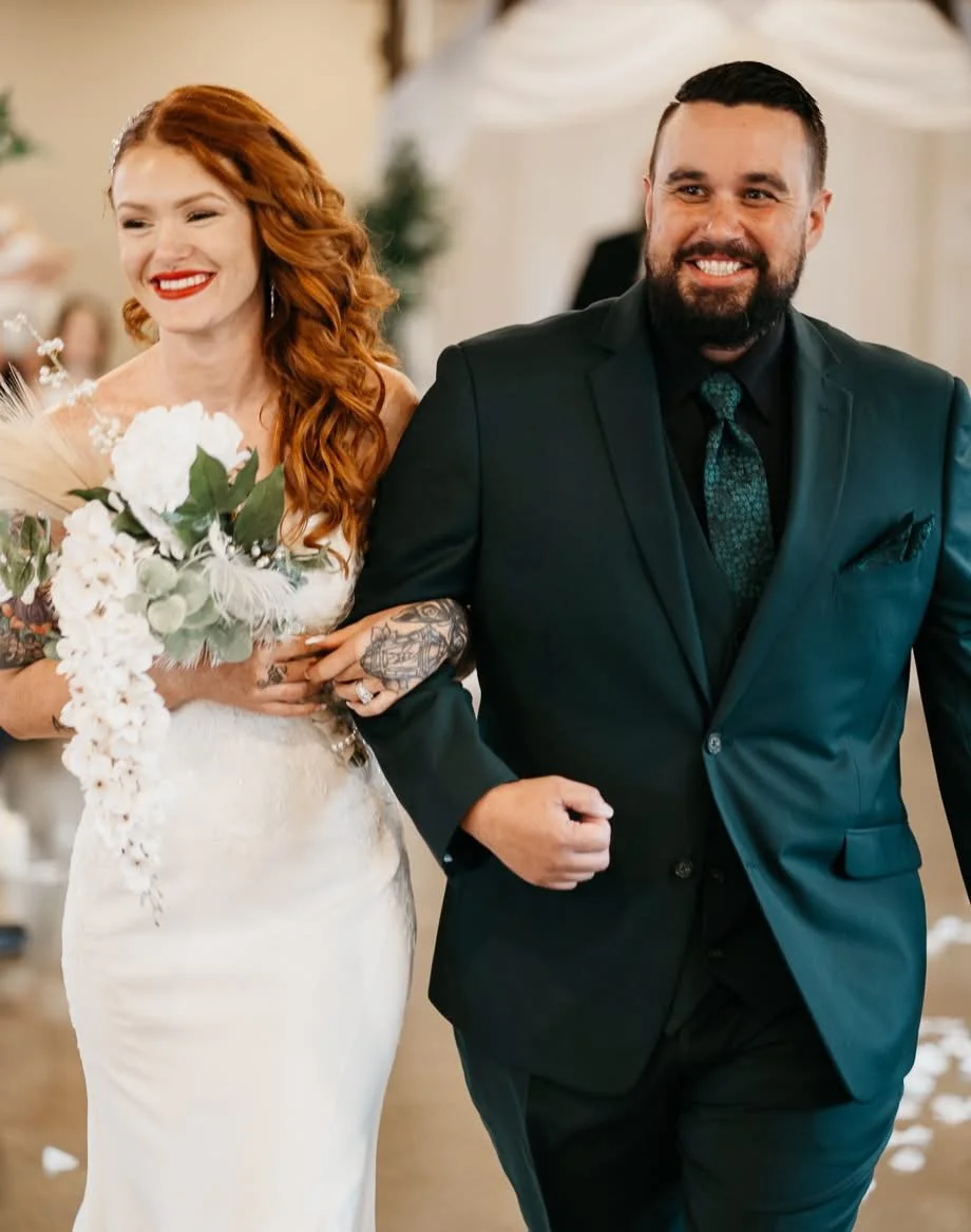 A smiling bride with red hair holding a bouquet, walking down the aisle with a groom with a beard, dressed in a dark suit, at a wedding ceremony.