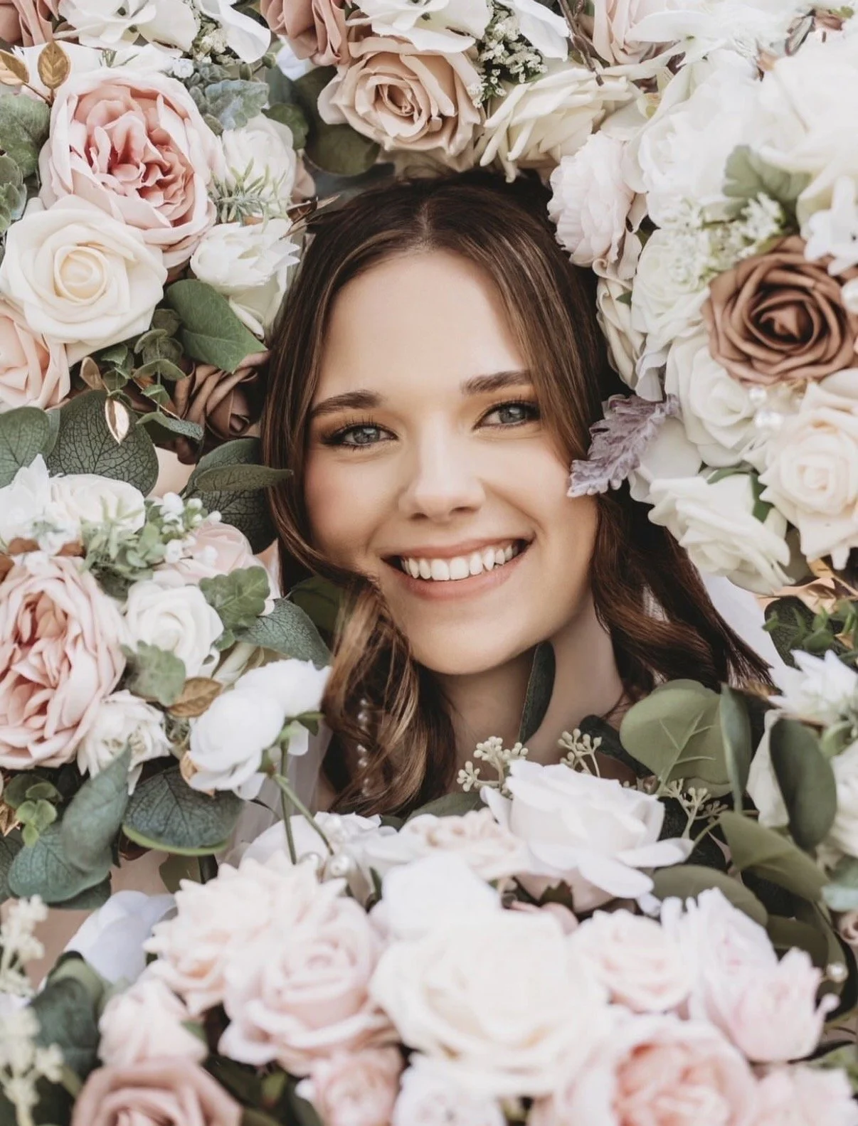 A woman with brown hair and blue eyes smiling among a collection of pink, white, and brown roses and green leaves.