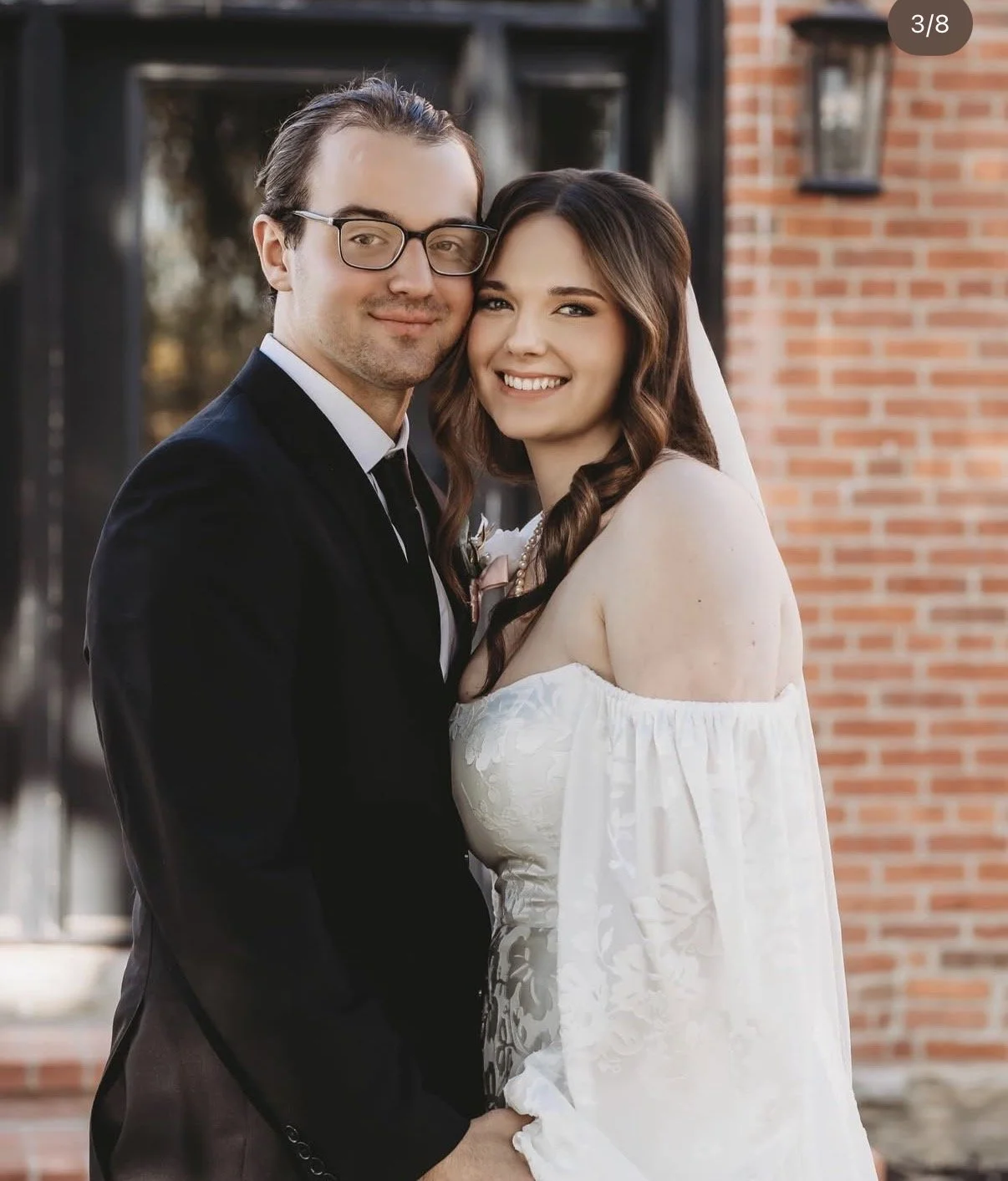 A young couple dressed in wedding attire, standing close together and smiling outdoors in front of a brick building.