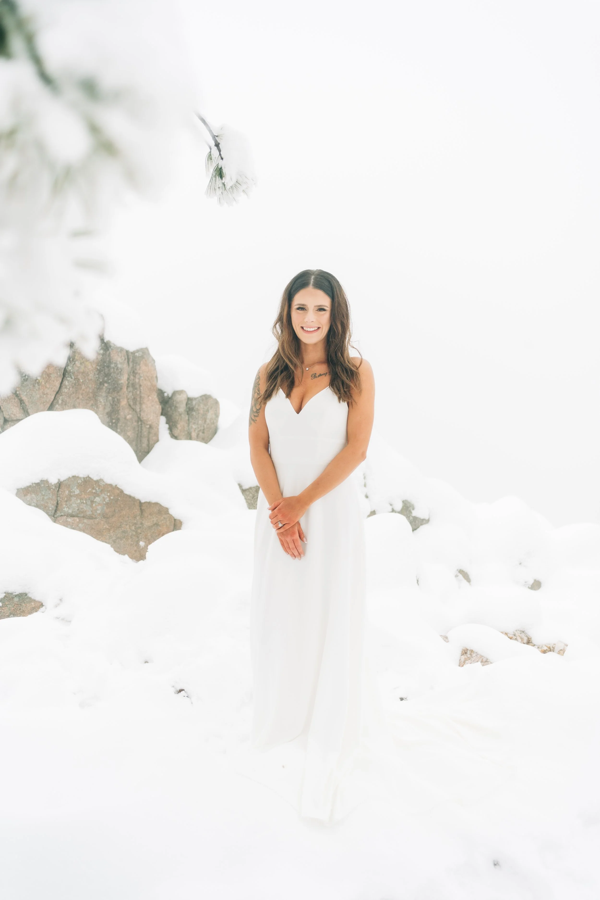 A woman in a white dress standing outdoors in a snowy landscape with rocks and trees in the background, smiling at the camera.