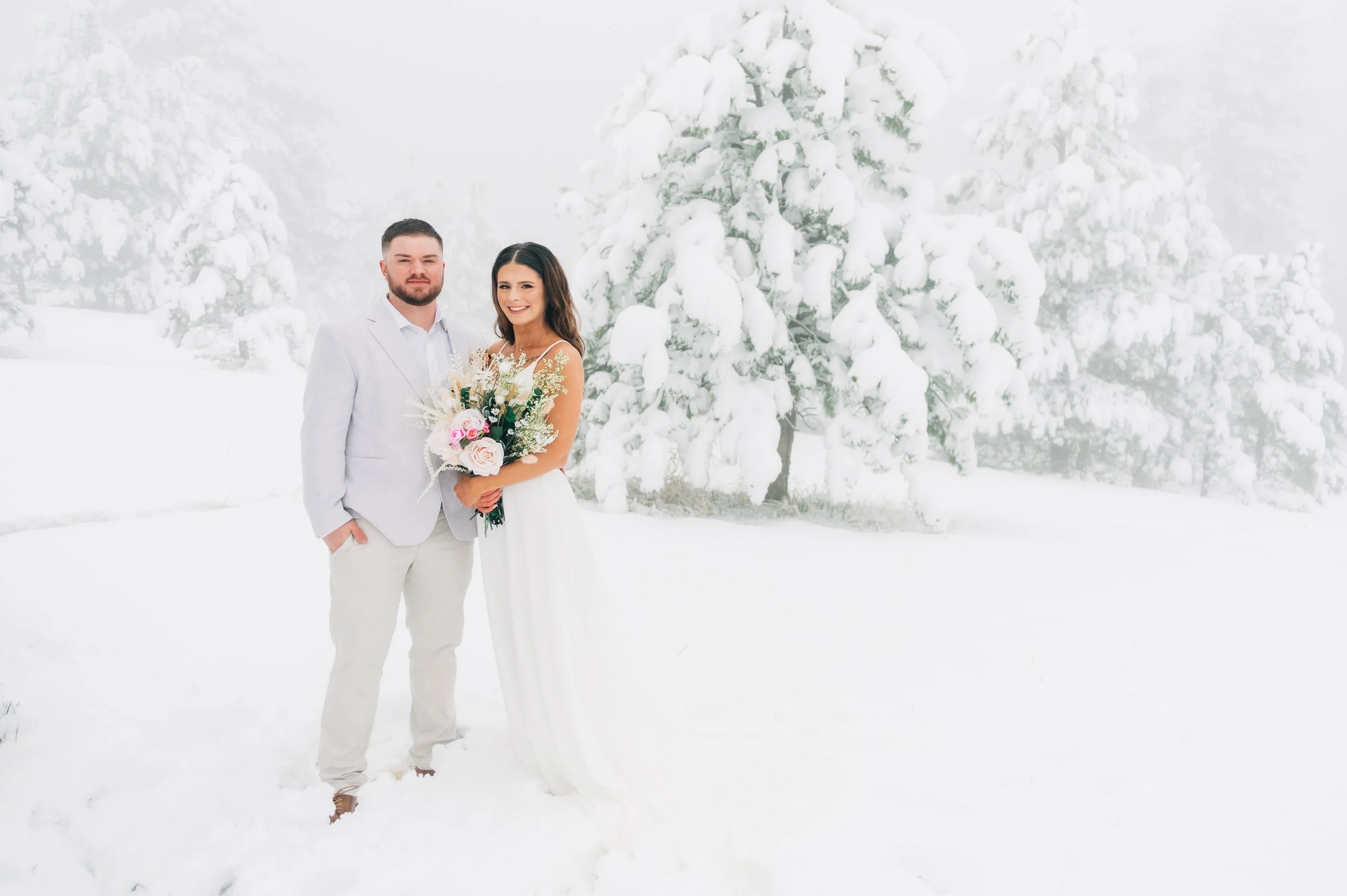 A couple in wedding attire standing in a snow-covered landscape with snow-laden trees in the background. The woman holds a bouquet of flowers and smiles, while the man stands beside her with his hand in his pocket.