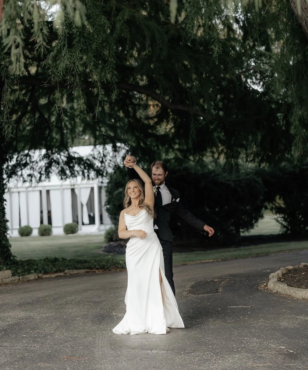 A bride holding a bouquet of greenery and white flowers, wearing a wedding dress and veil, smiling near a window.