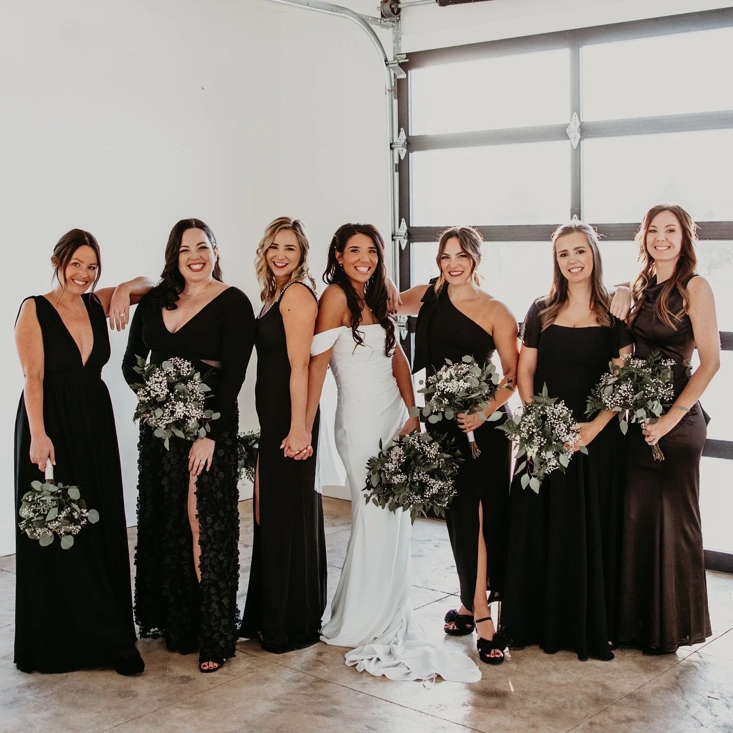 A group of eight women, dressed in black and white formal dresses, standing in a bright indoor space with a garage door window in the background, holding bouquets of flowers, smiling for a photo.