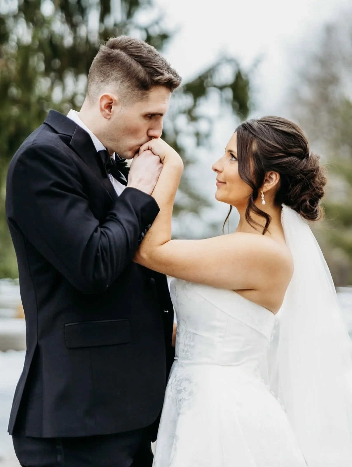 A bride holding a bouquet of white roses and greenery, smiling with her eyes closed outdoors.