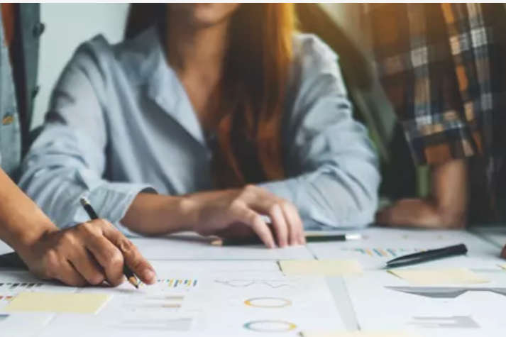 A woman with short blonde hair, wearing an orange sleeveless top and beige pants, sitting on a beige cushion at a modern indoor space, writing in a notebook. Behind her is a large green plant, a wooden panel, and abstract art with shadowed plant imagery on the wall, illuminated by natural sunlight.