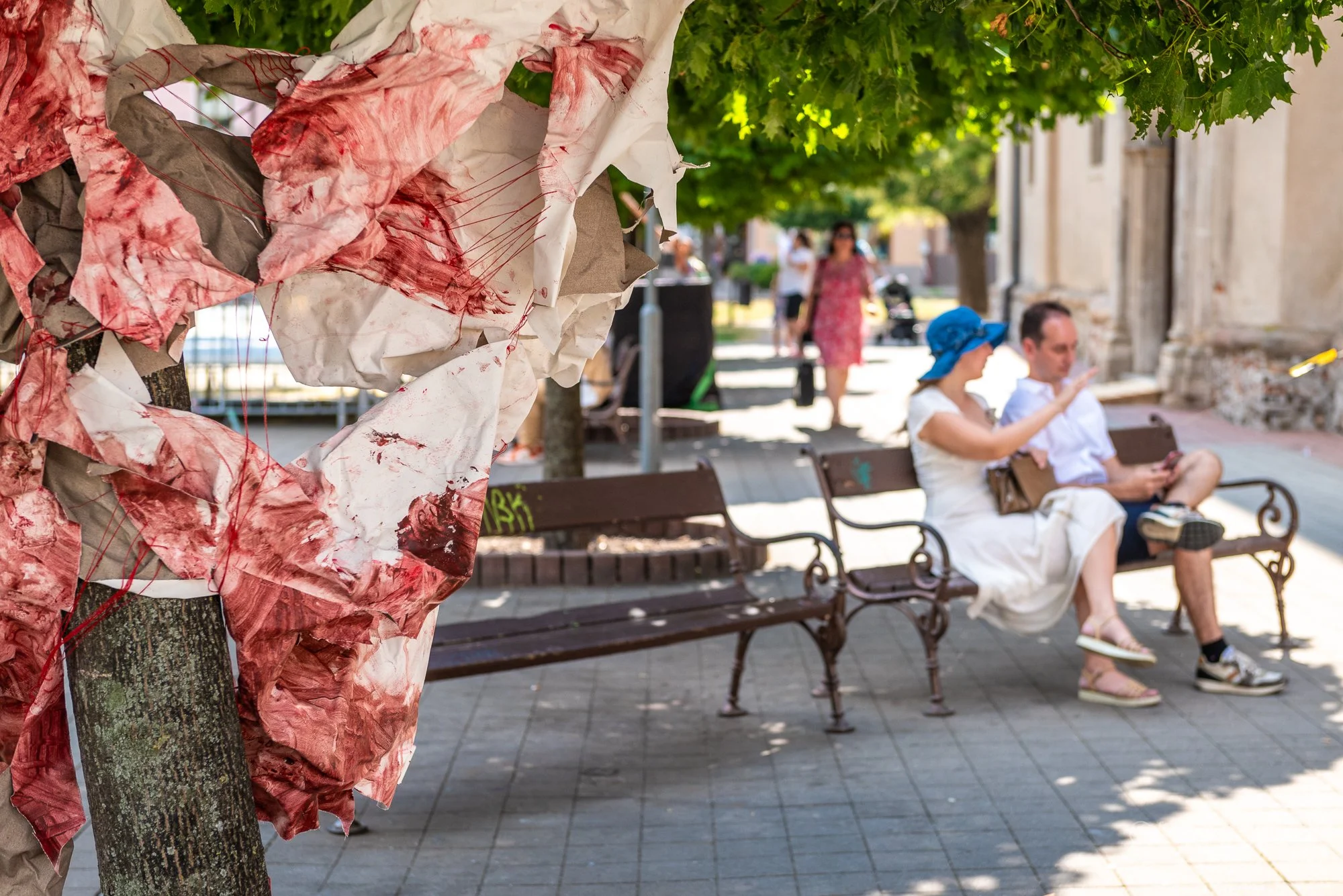 A close-up of a crumpled, pink and white paper decoration attached to a tree trunk in the foreground. In the background, a woman in a white dress and a blue sun hat and a man in a white shirt and shorts sit on a park bench, with the woman looking at her phone and the man also focused on his phone. Further back, a woman in a pink floral dress walks along a pathway shaded by green trees, with more people and buildings visible in the distance.