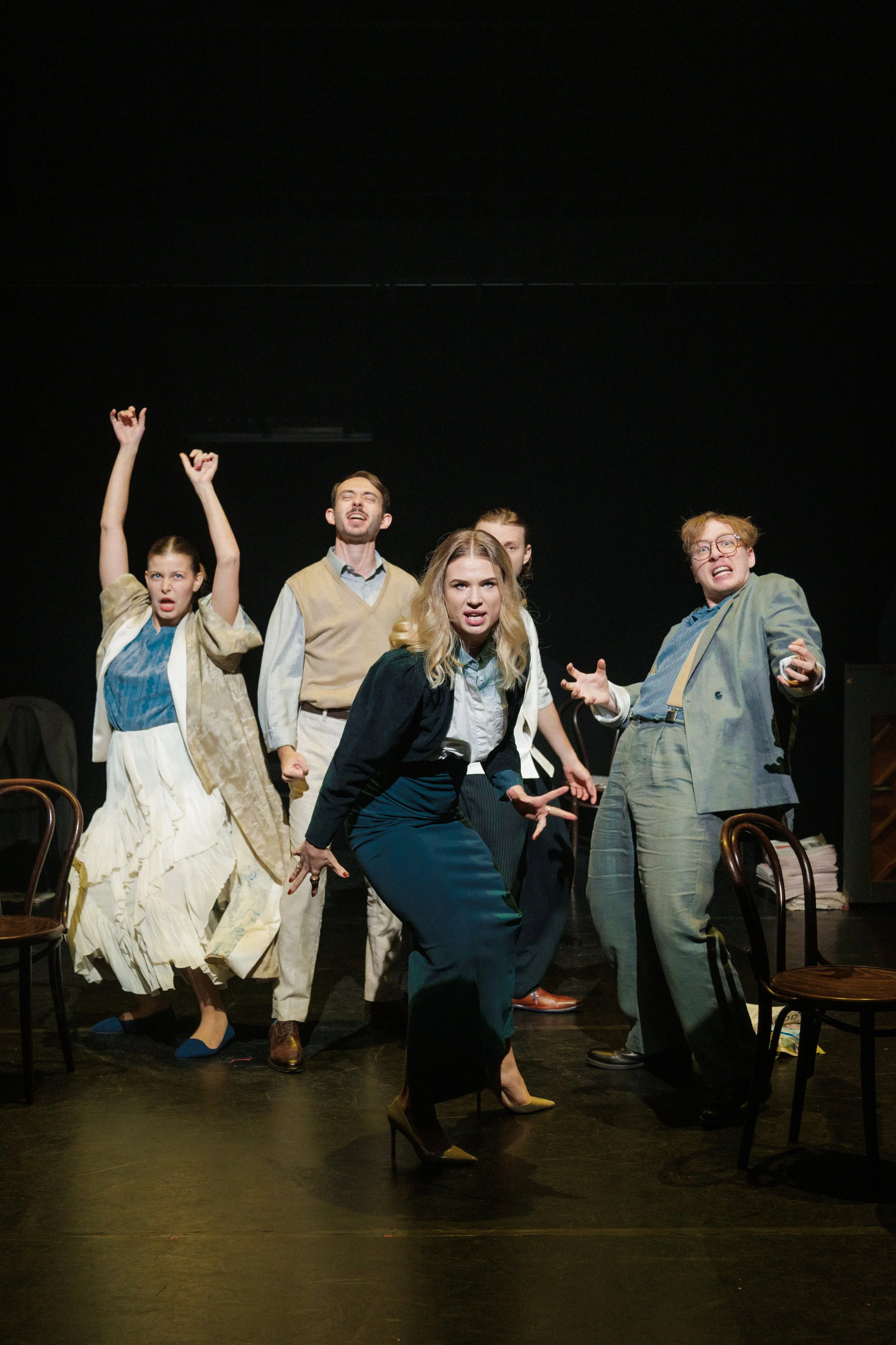 Group of actors performing on stage with expressions and gestures, some seated and some standing.