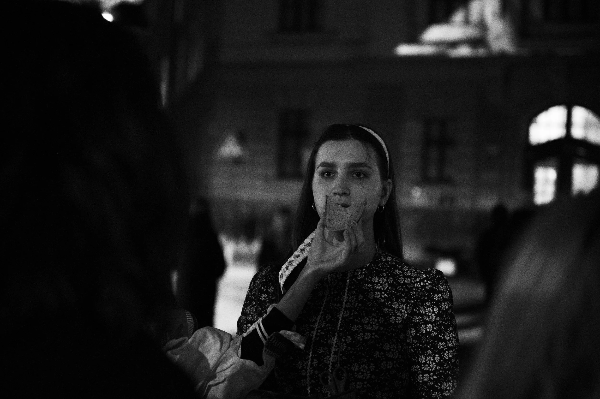 A young woman with long hair and earrings is holding a slice of bread near her mouth inside a dimly-lit wooden interior with windows in the background.