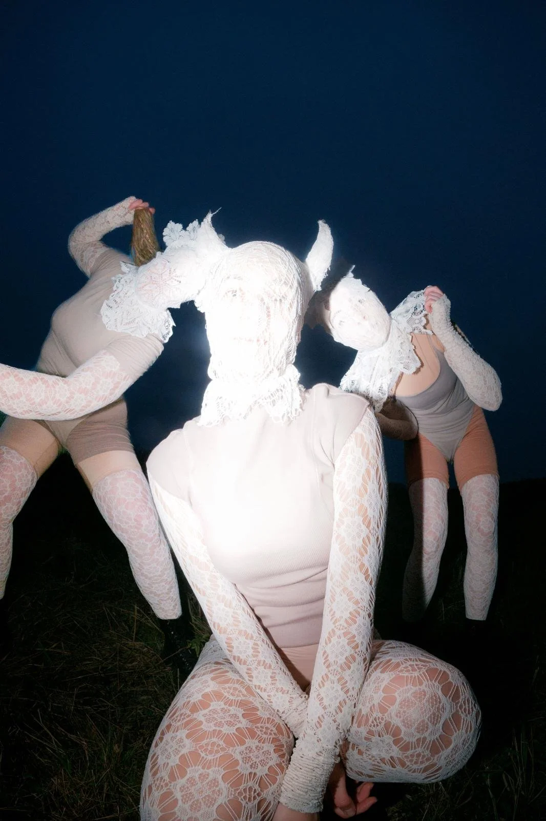 Three individuals dressed in lace and spaghetti straps, with their faces covered by lace masks, holding objects in a dark outdoor setting at night.