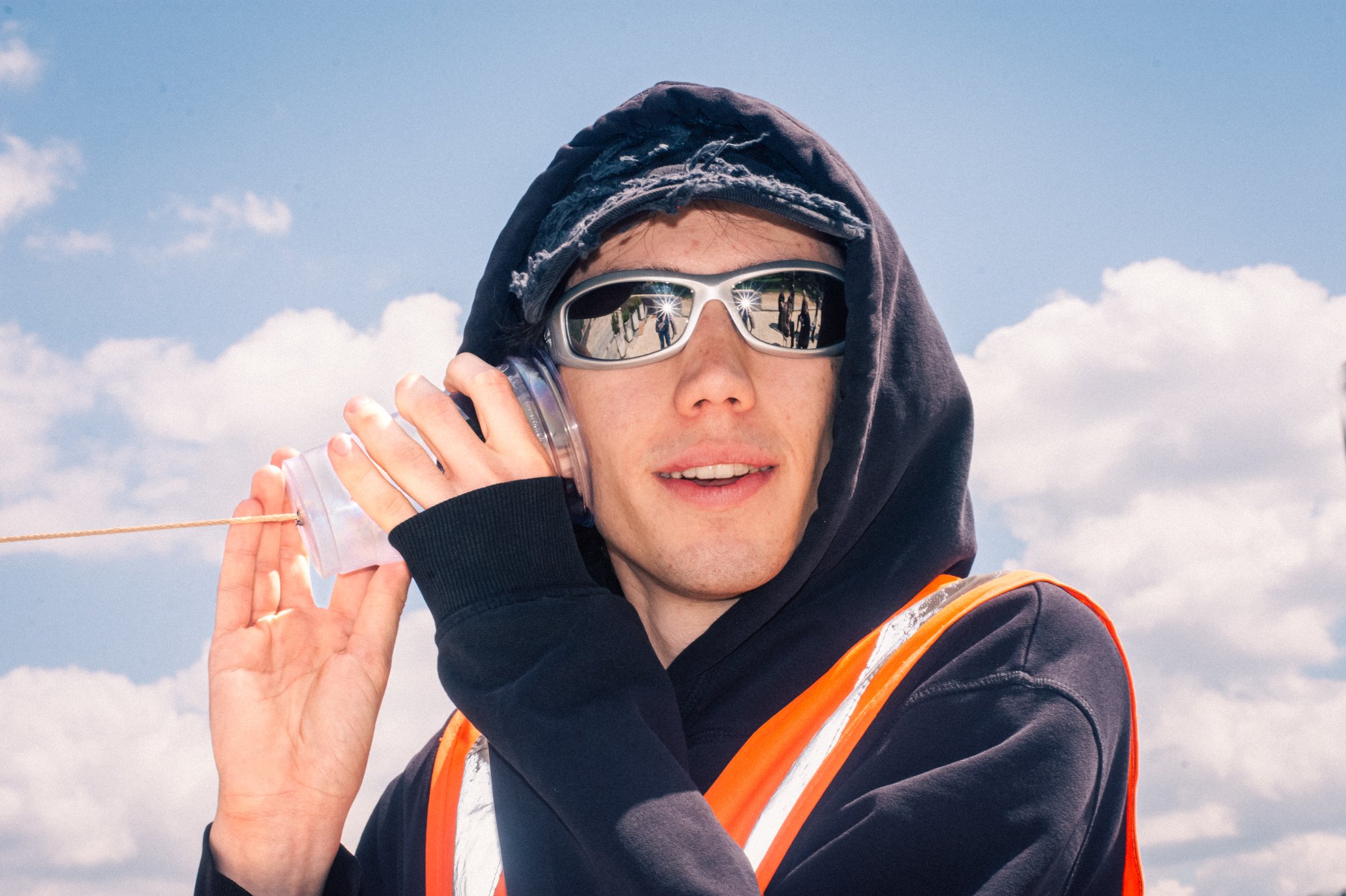 A young man wearing sunglasses and a hooded jacket holding a transparent water bottle to his ear outdoors under a partly cloudy sky.