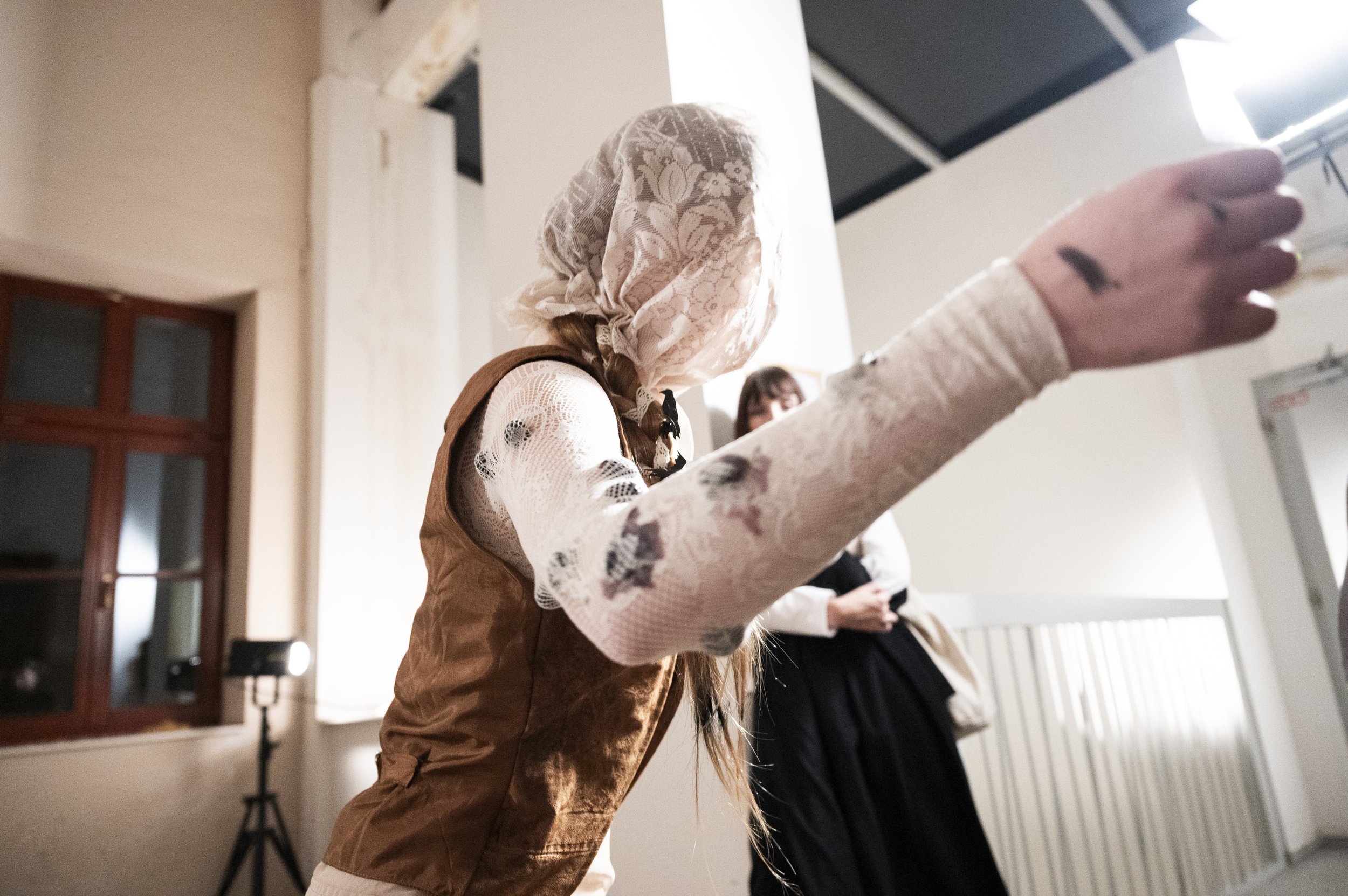 A person wearing a lace headscarf and a lace shirt is raising one arm. Another person in a black dress is standing in the background in an indoor setting.