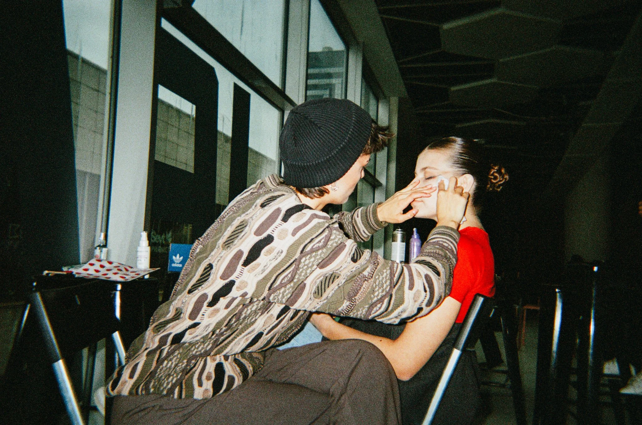 A person applying makeup to another person sitting on a chair in a modern indoor setting.