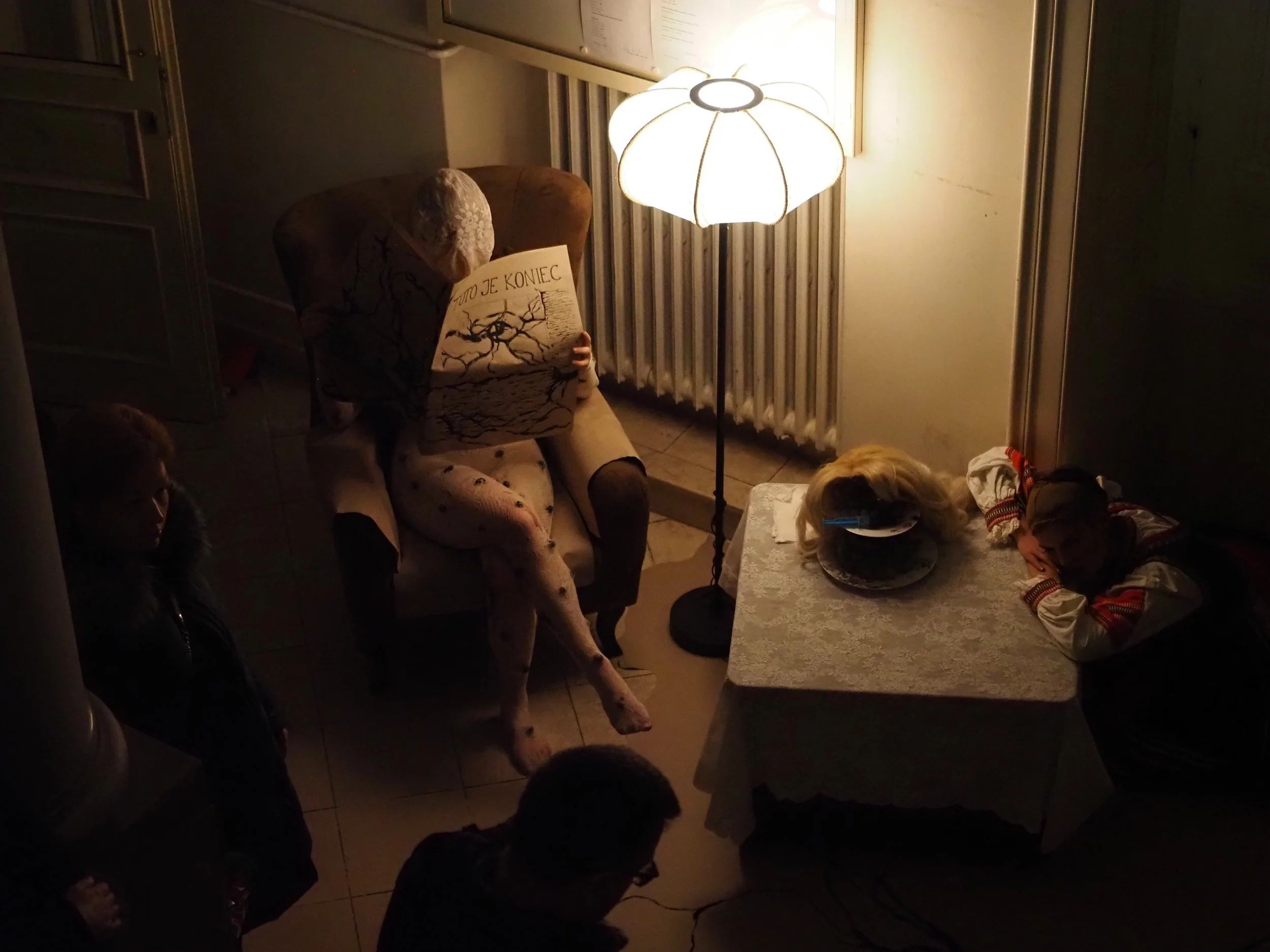 A person with a head covering sitting in a brown armchair reading a book, illuminated by a floor lamp, with a small table next to them. Two people are lying on the floor, one on a table, in a dimly lit room with a radiator and patterned wallpaper.