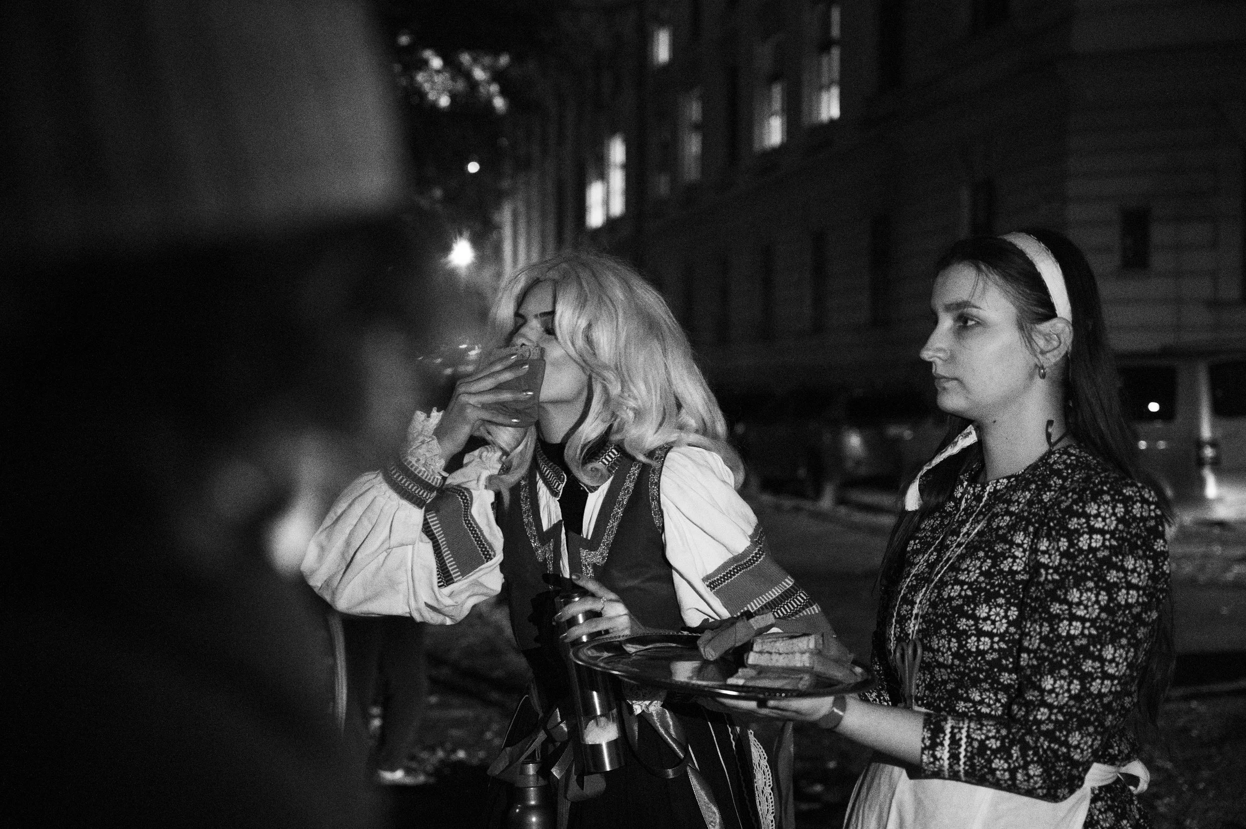 Two women standing outdoors at night, one is drinking from a cup and the other is holding a tray with drinks, in front of a background of tall buildings.