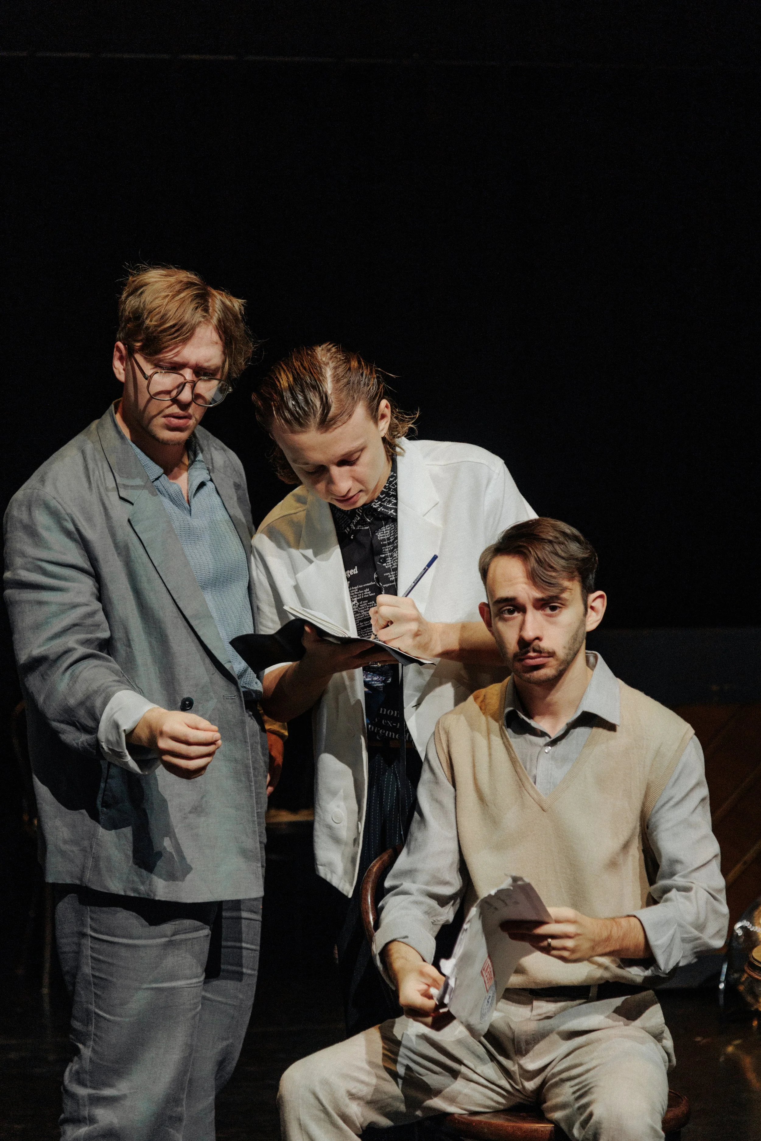 Three actors on stage, two men standing and one man sitting, engaged in a scene with serious expressions, against a plain dark background.