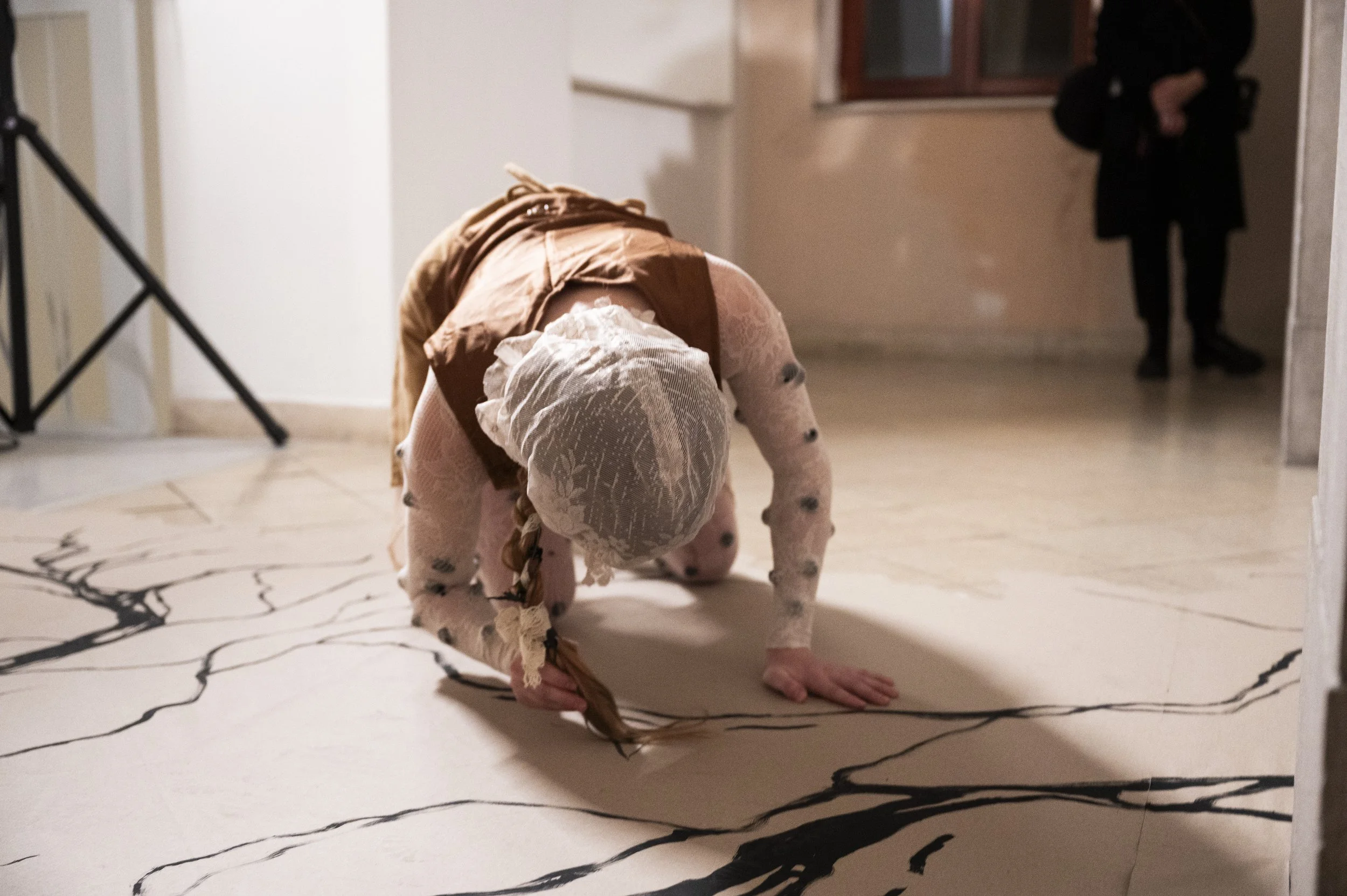 A person dressed in a lace outfit with a head covering is crawling on the floor in an indoor space with a patterned floor. In the background, a person dressed in black stands near a window.