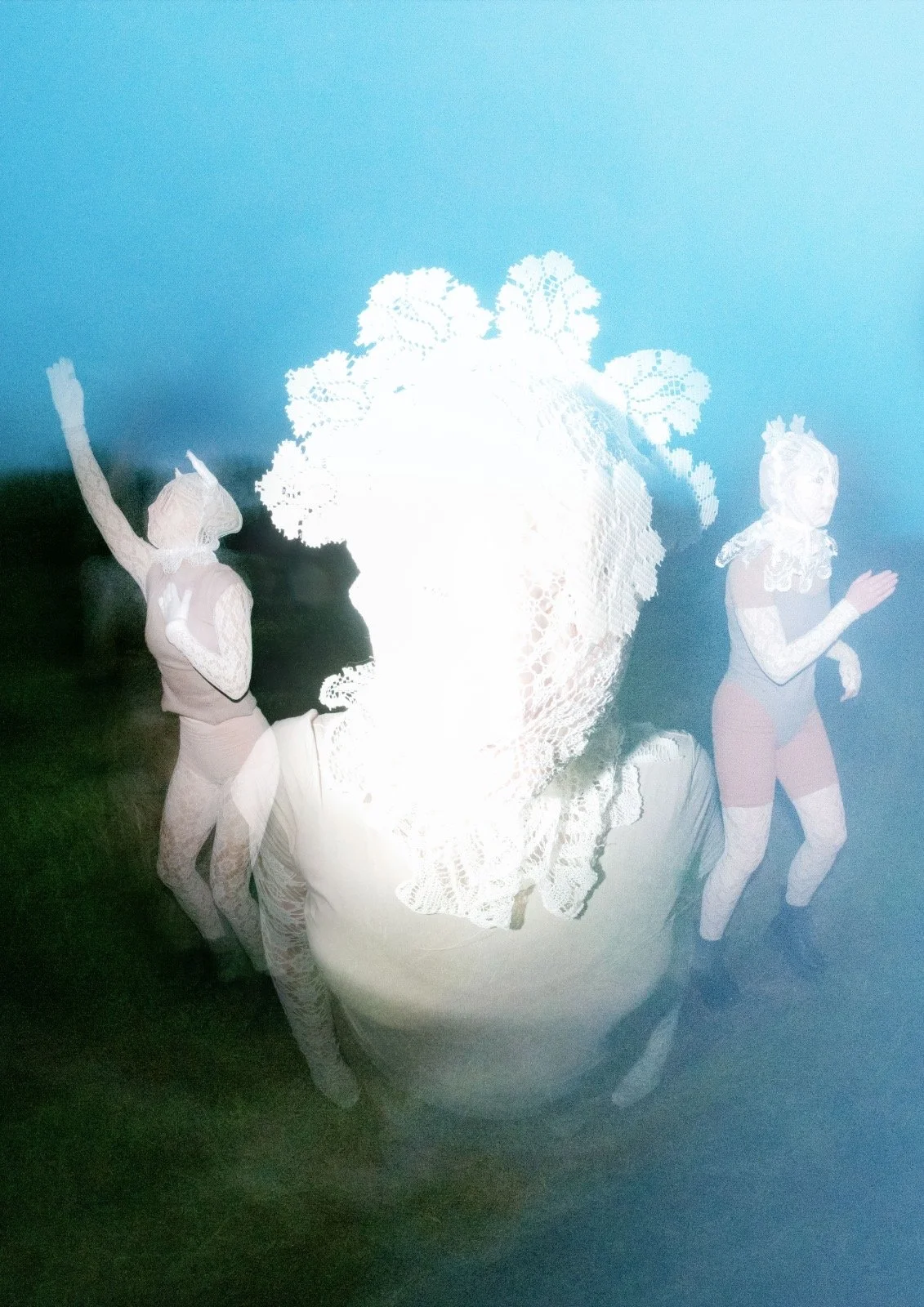 A blurred photo of three people in retro-style costumes outdoors during twilight; two are women with short hair, one raising an arm, the other walking, wearing lace-like tops and shorts, while the third person has a large, detailed, lace collar obscuring their face.