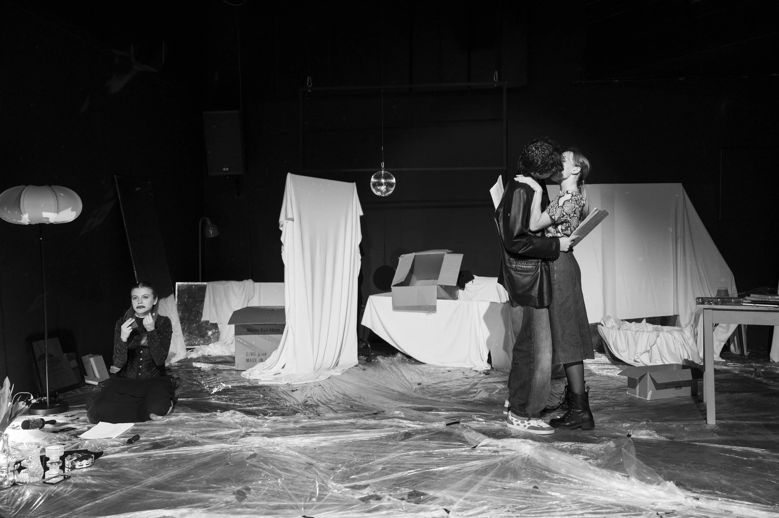 Two people kissing on stage, while a woman sits on the floor nearby. The stage has miscellaneous props, boxes, and drapes, with a plastic floor covering and a disco ball hanging from above.