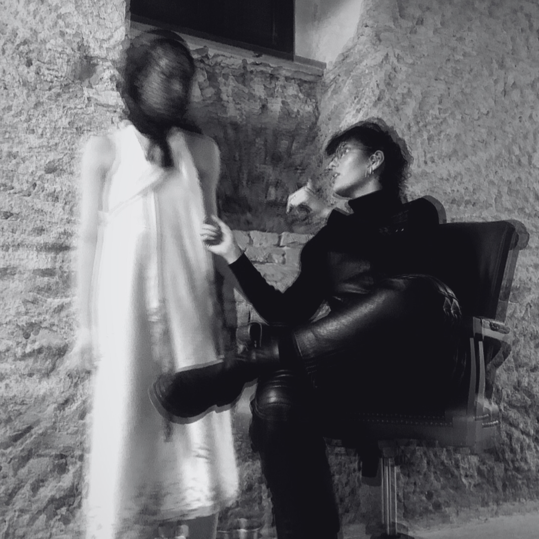 A woman seated in a chair holding another woman's hand inside a room with stone walls, photographed in black and white.