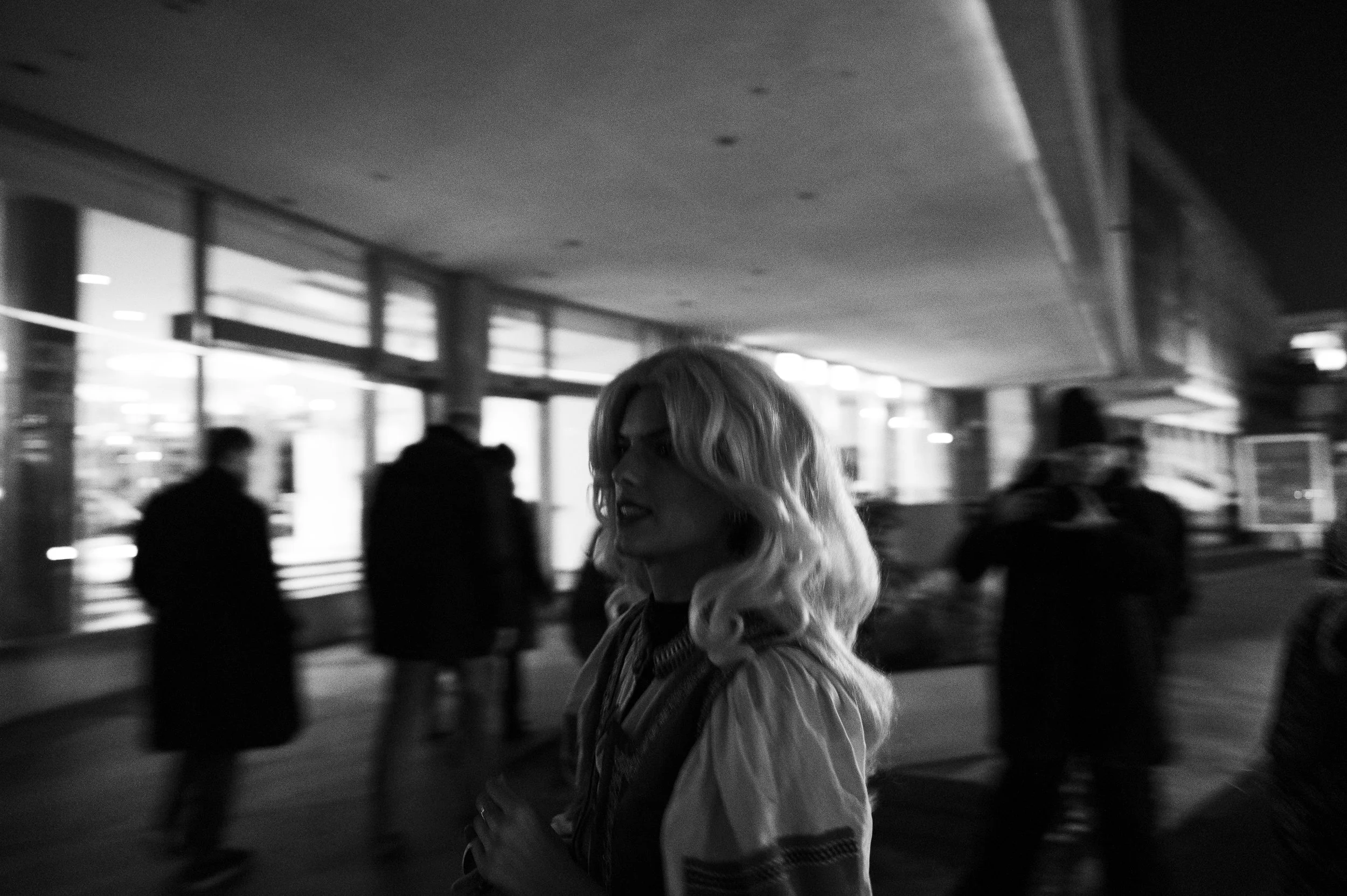 A black and white photo of a woman with wavy blonde hair standing in a busy urban area at night, with blurred people walking past and storefronts illuminated behind her.