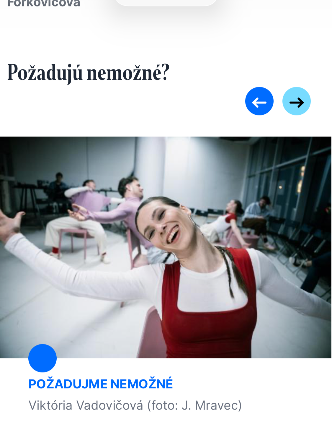 A group of people sitting on chairs in a room with large windows, engaging in a performance or dance, with a smiling woman in front stretching her arms.