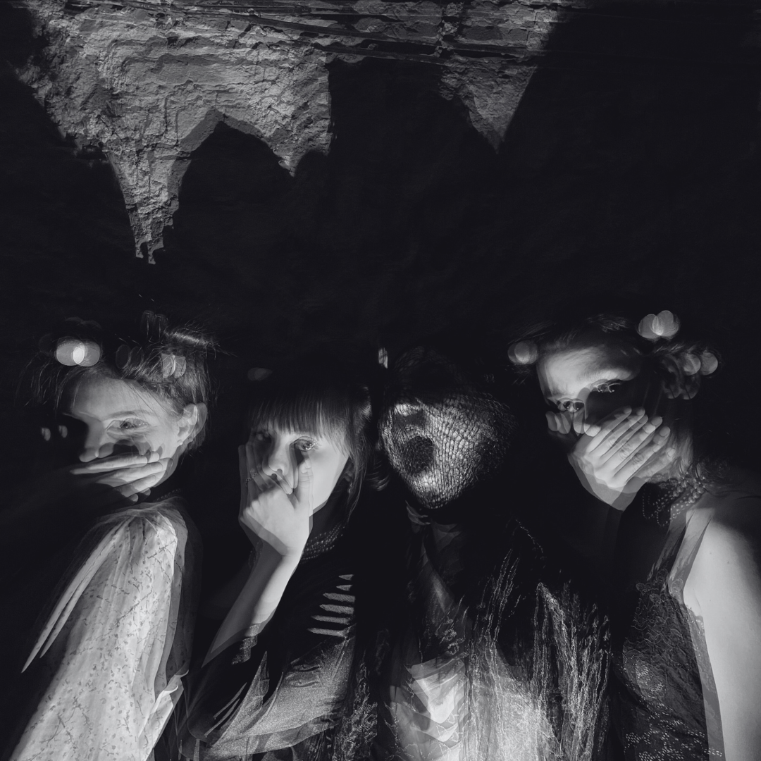 Four women in dark clothing with varied hairstyles posing closely together in a dark setting, with a textured ceiling or rock formation overhead and light reflections creating a bokeh effect.