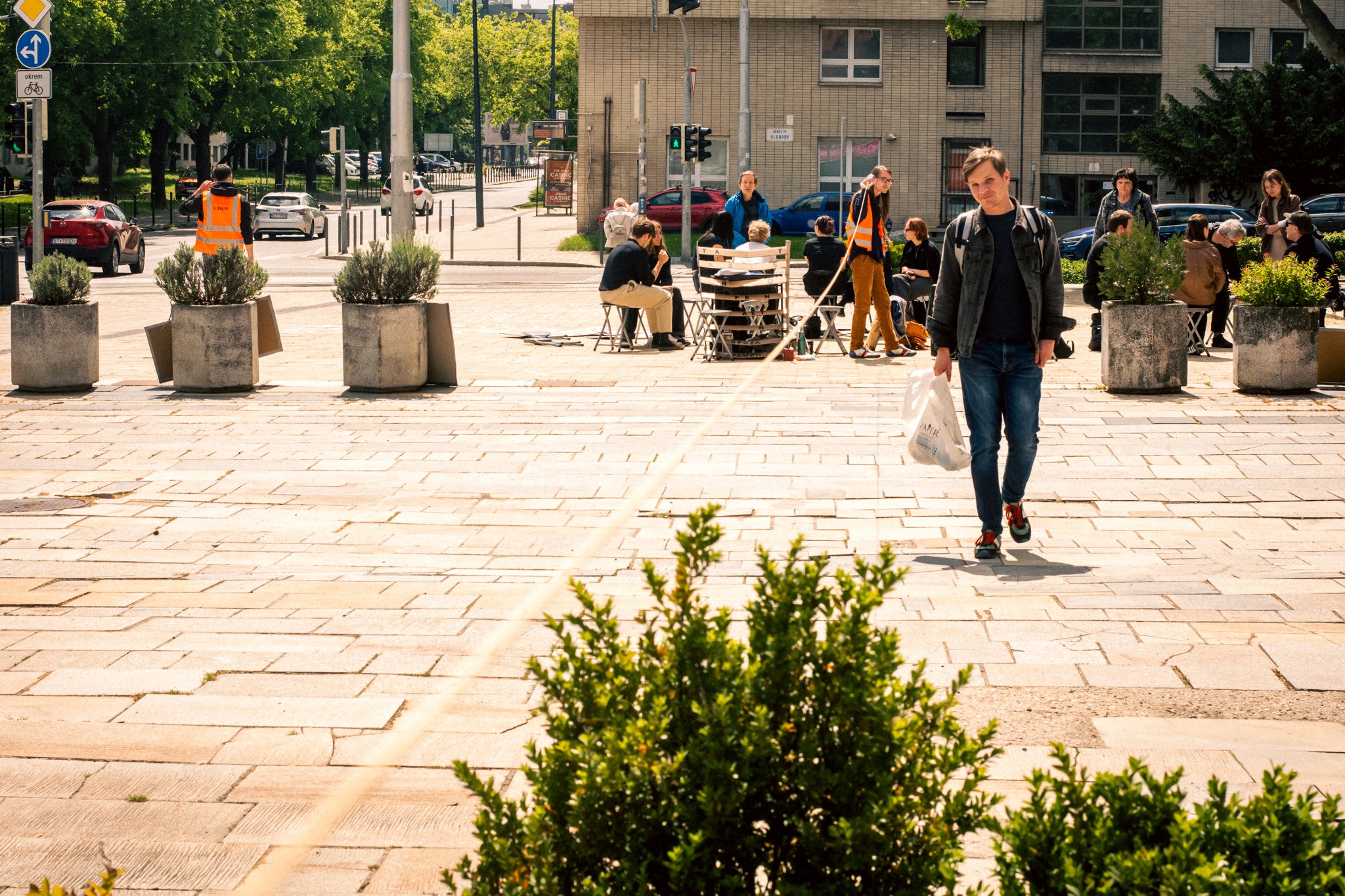 A young man walking on a city sidewalk, carrying a shopping bag, with a group of people seated and standing in the background near a train crossing signal, trees, and buildings.