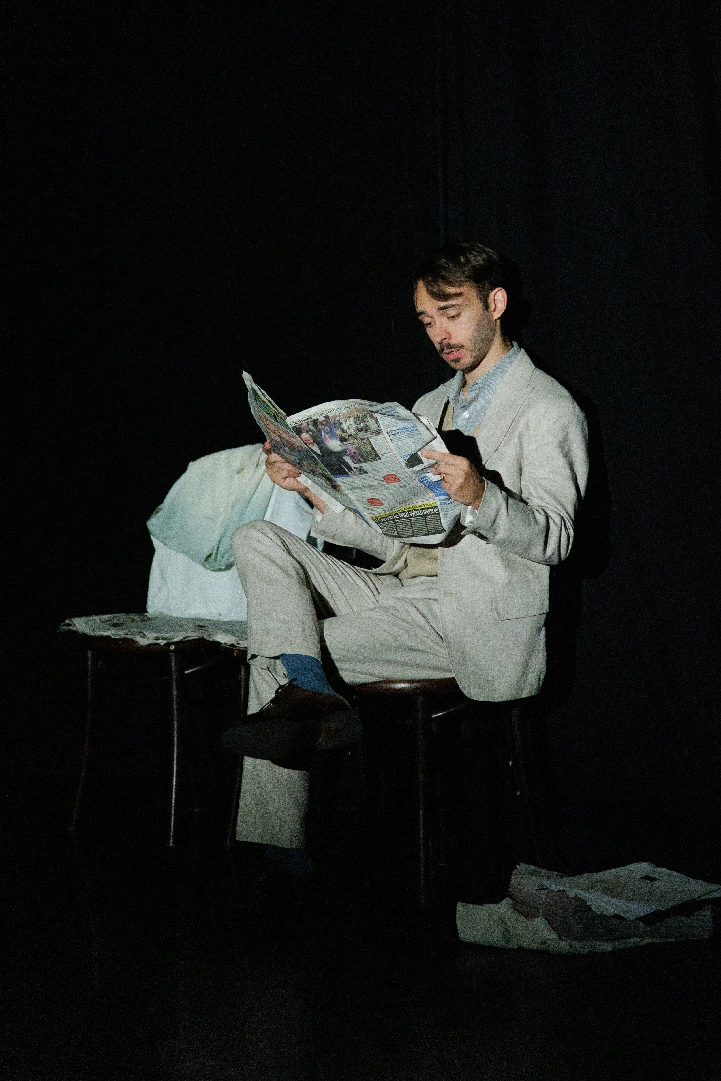 A man in a light gray suit sitting on a chair, reading a newspaper with a dark background.