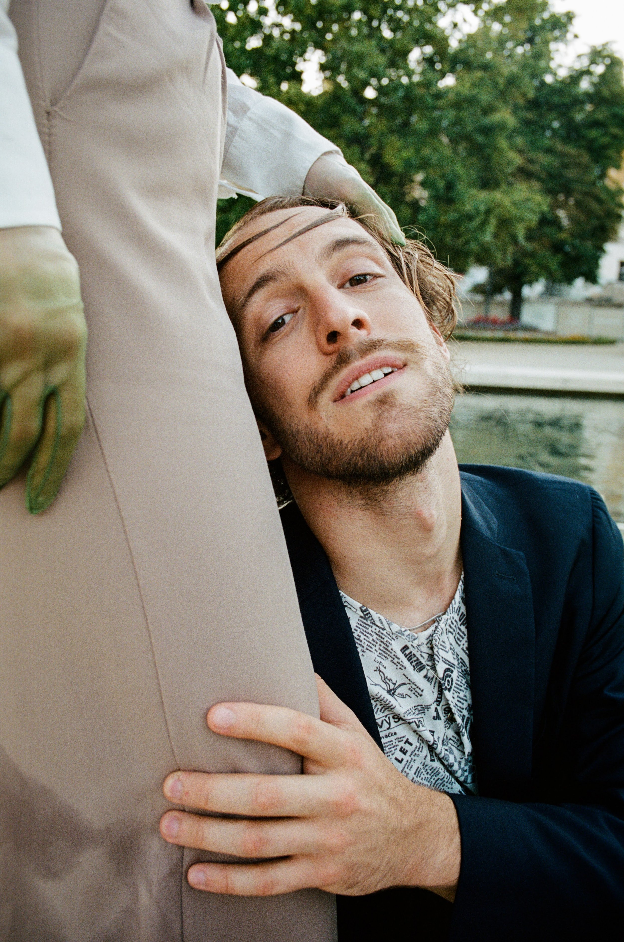 A man with a beard and wavy hair is kneeling by a riverbank, resting his head against a person's leg, who is standing and wearing beige pants and gloves. Trees and a water body are visible in the background.