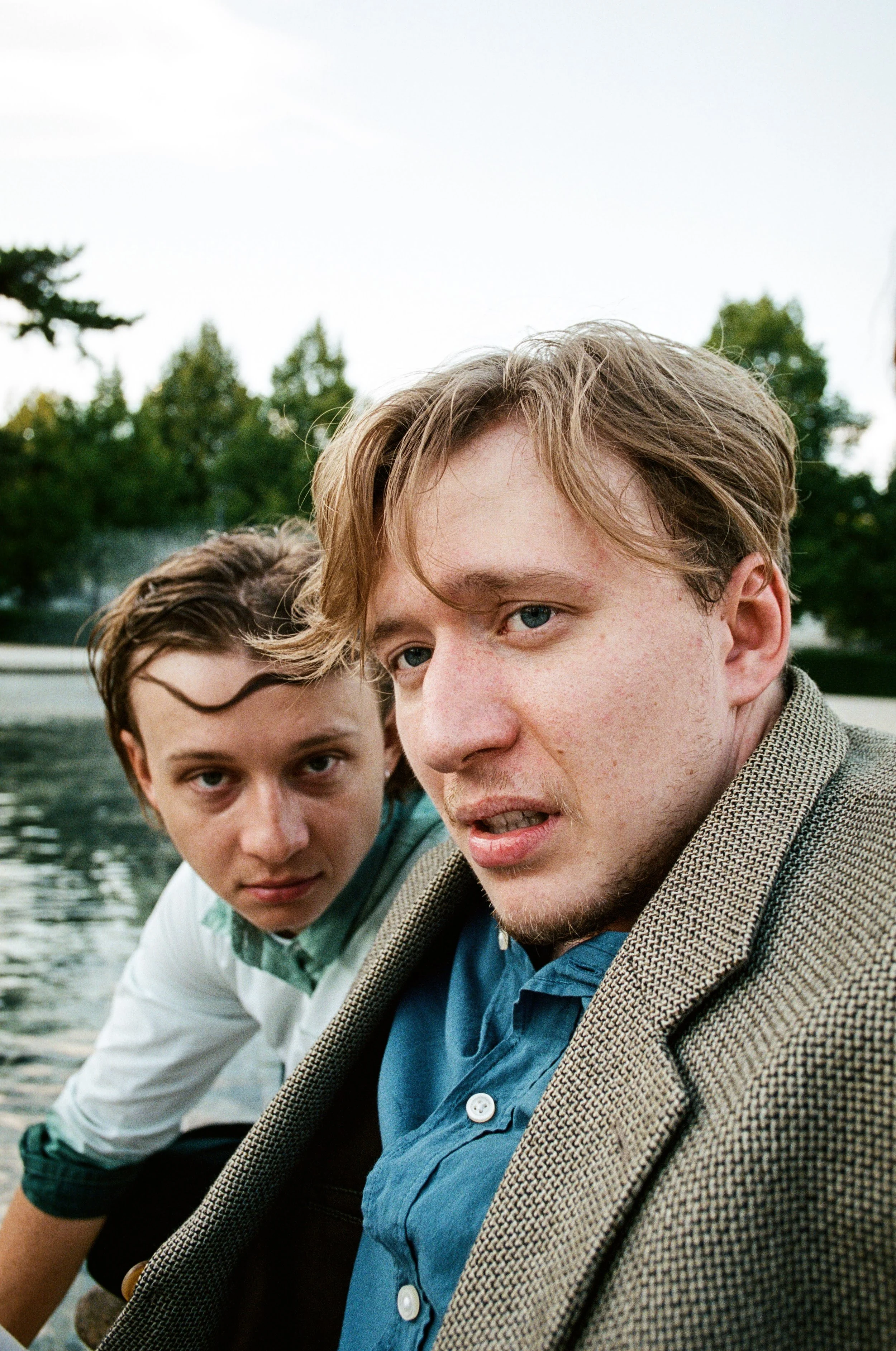 Two young men sitting by a body of water outdoors, with trees in the background. One has light brown hair and the other dark brown hair, both looking toward the camera with serious expressions.