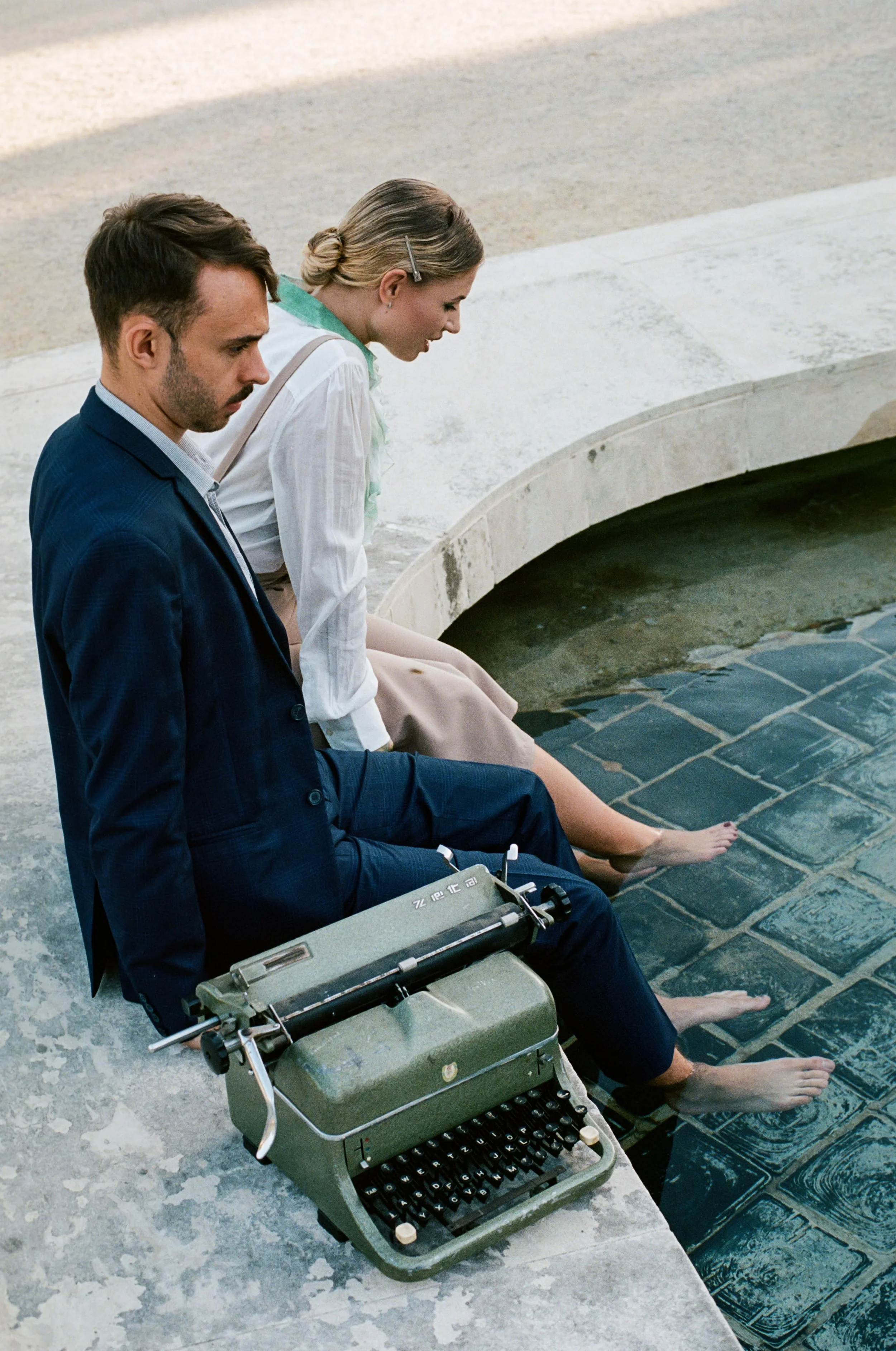 A man and a woman sitting at the edge of a fountain with their feet in the water, and a vintage typewriter placed on the ground beside them.