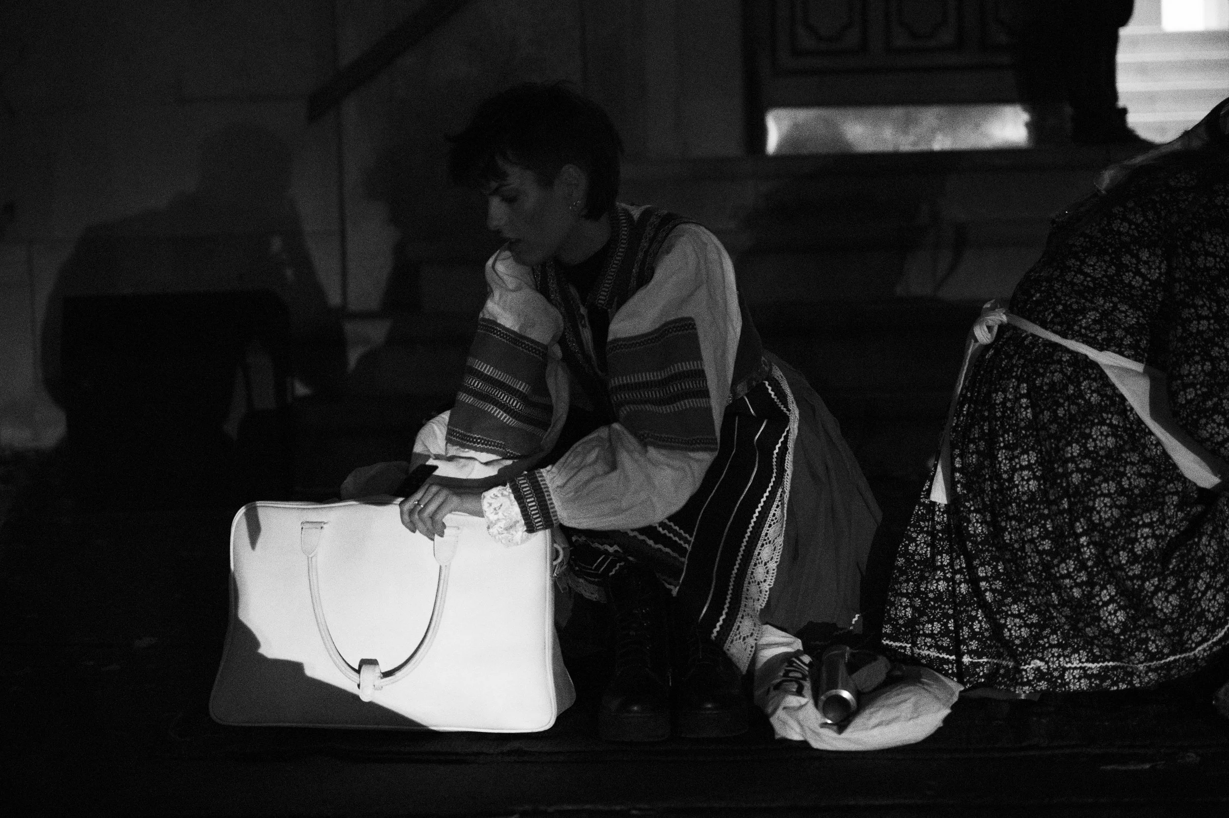A woman dressed in traditional clothing sitting on the floor with a large white bag in front of her, in a dimly lit indoor setting.