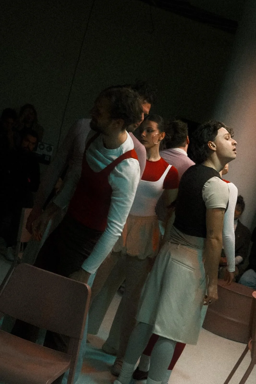 A group of people standing in a dimly lit room, with some individuals wearing retro-style clothing, possibly during a performance or event.