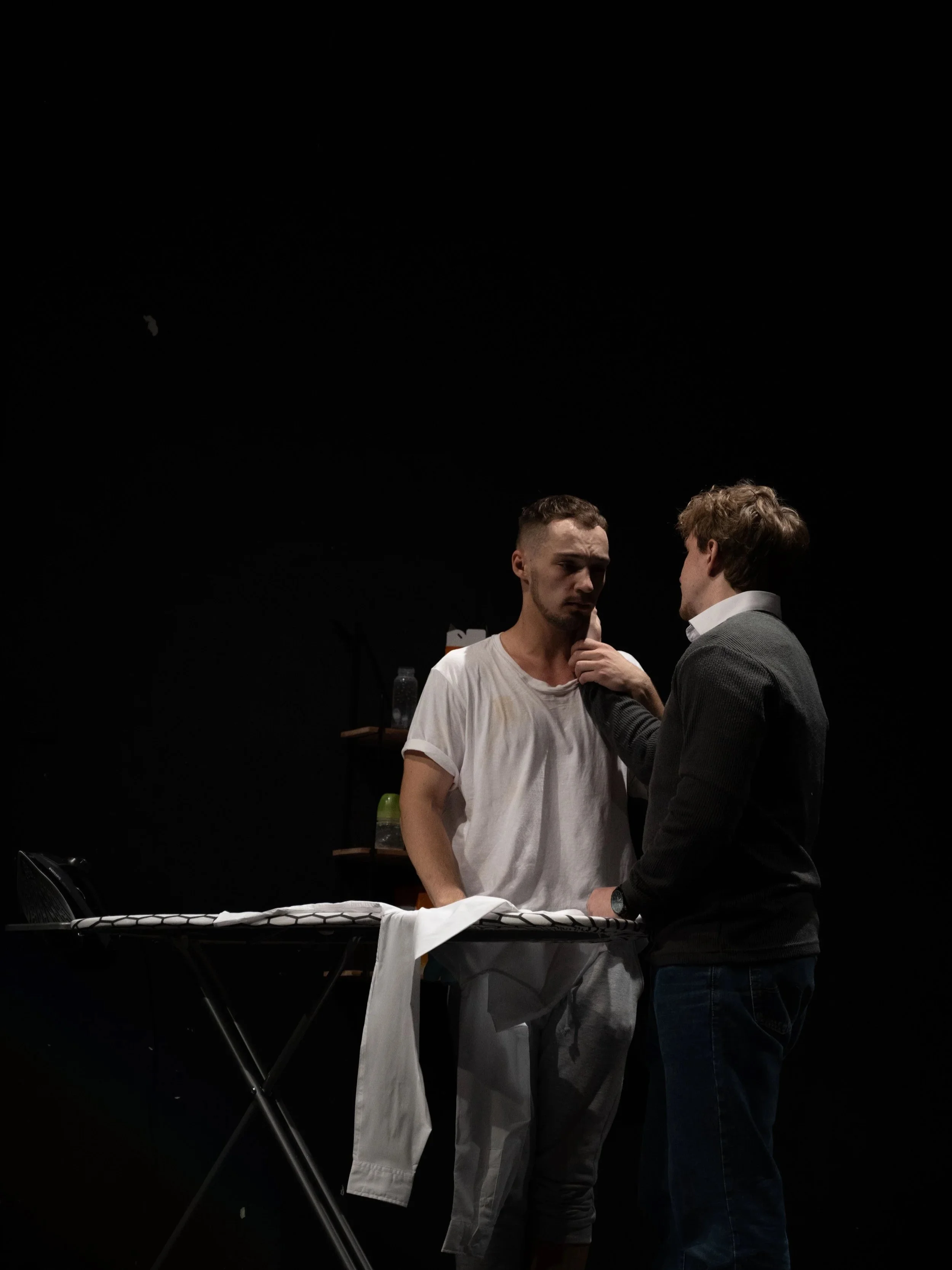 Two young men are engaged in a serious conversation in a dimly lit room with a black background. One man is wearing a white t-shirt and grey sweatpants, while the other is dressed in a dark sweater and jeans. An ironing board with clothes draped over it and some bottles in the background are visible.