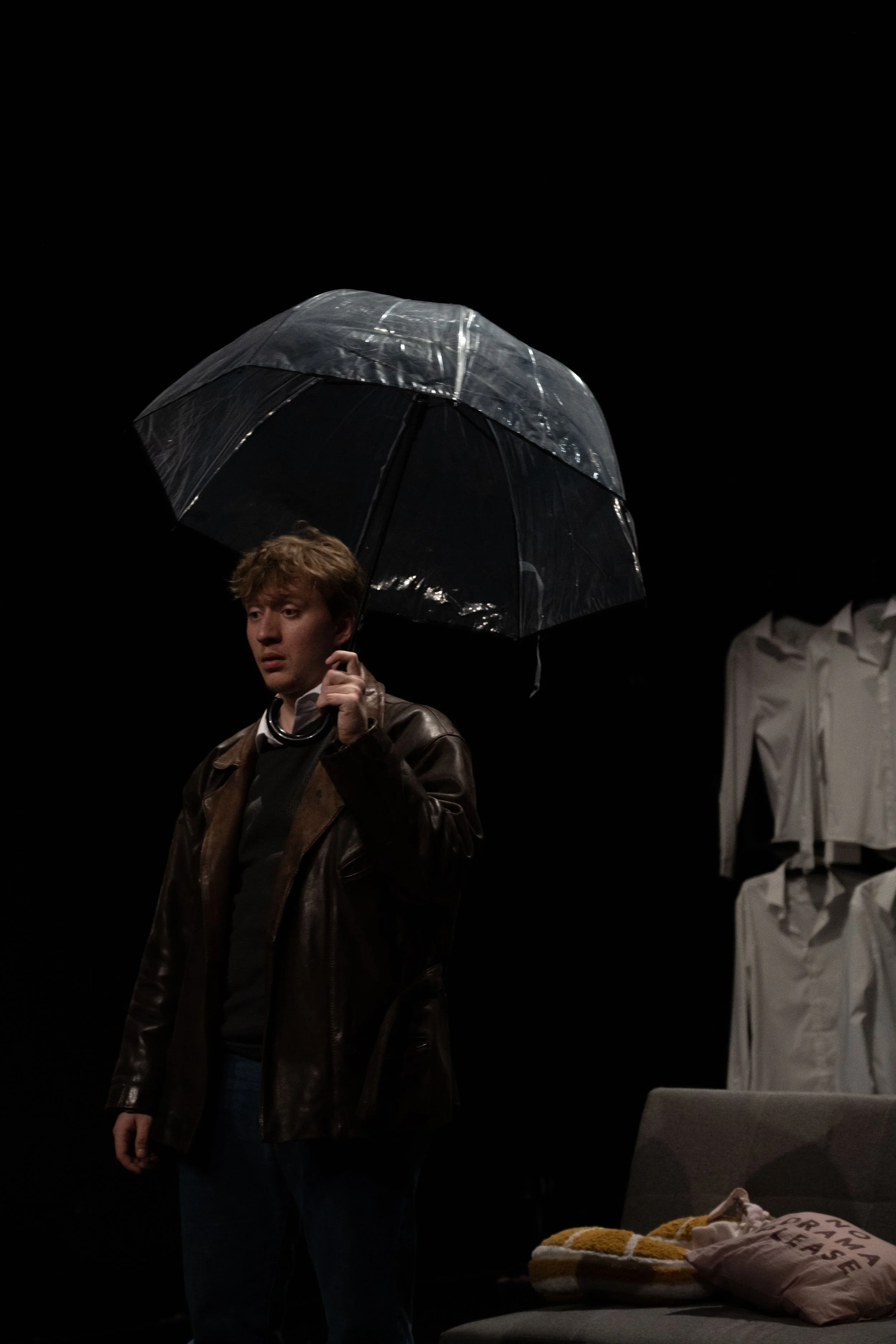 A young man holding a black umbrella on a dark stage, dressed in a brown leather jacket and dark pants, standing near a gray sofa with yellow and white pillows, with white draped fabrics in the background.