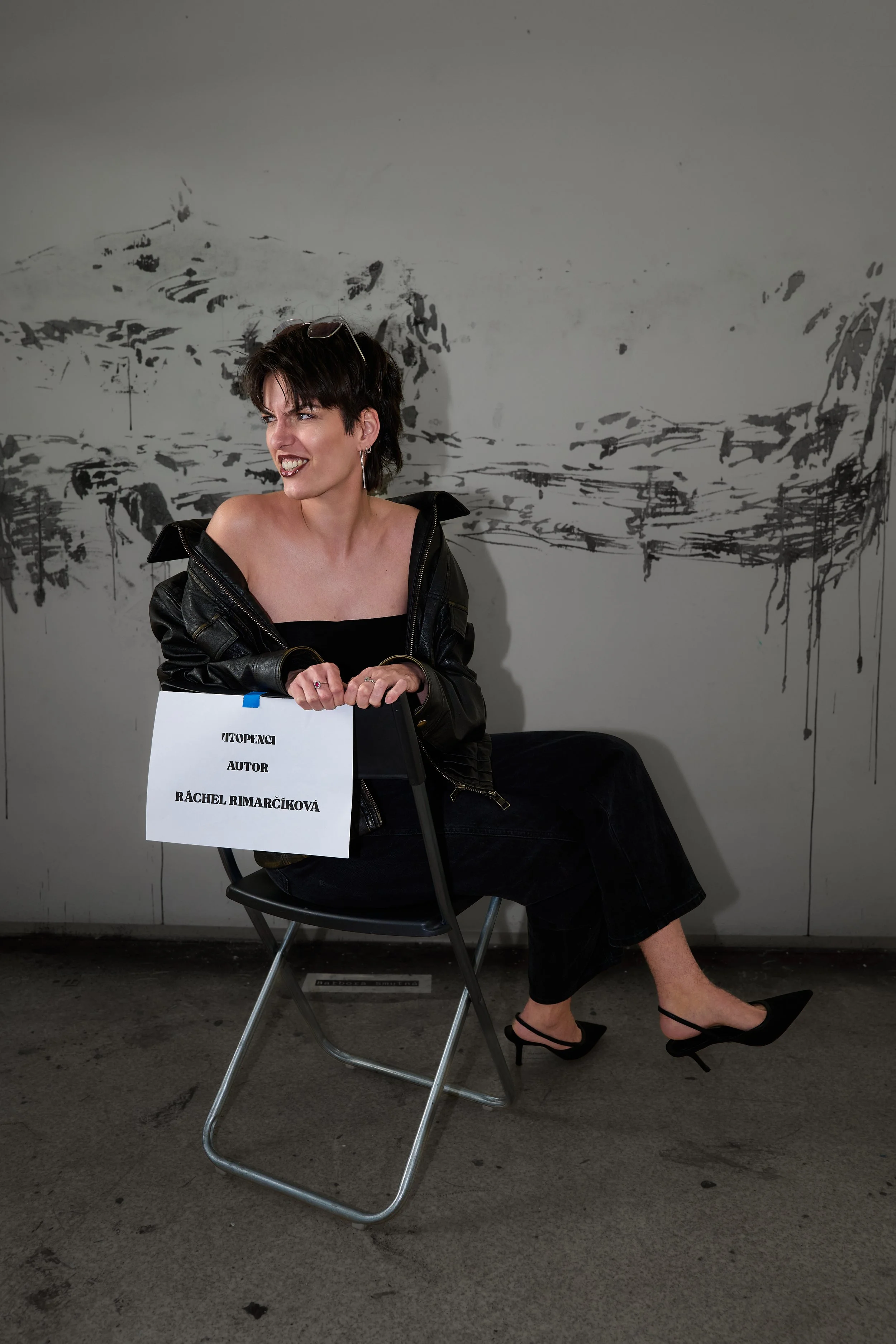 A woman with short dark hair, wearing a black dress and a black leather jacket, seated on a gray folding chair holding a sign. The sign reads 'UPOČNI AUTOR RÁCHEL RIMARČÍKOVÁ'. She is smiling slightly and appears to be in an indoor space with a gray wall featuring black abstract paint splatters in the background. She is also wearing black high-heeled shoes with ankle straps.