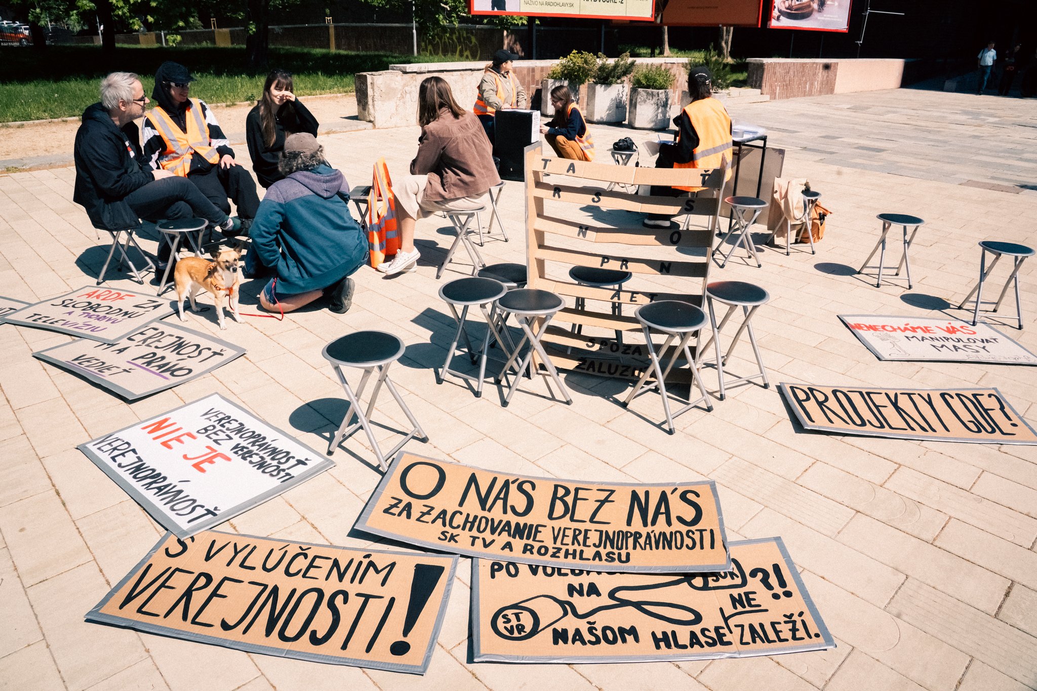 A group of people gathered outdoors, sitting and talking, with protest signs laid on the ground. Signs contain messages in Slovak about public service and honesty, and there are small stools and a wooden barrier nearby.