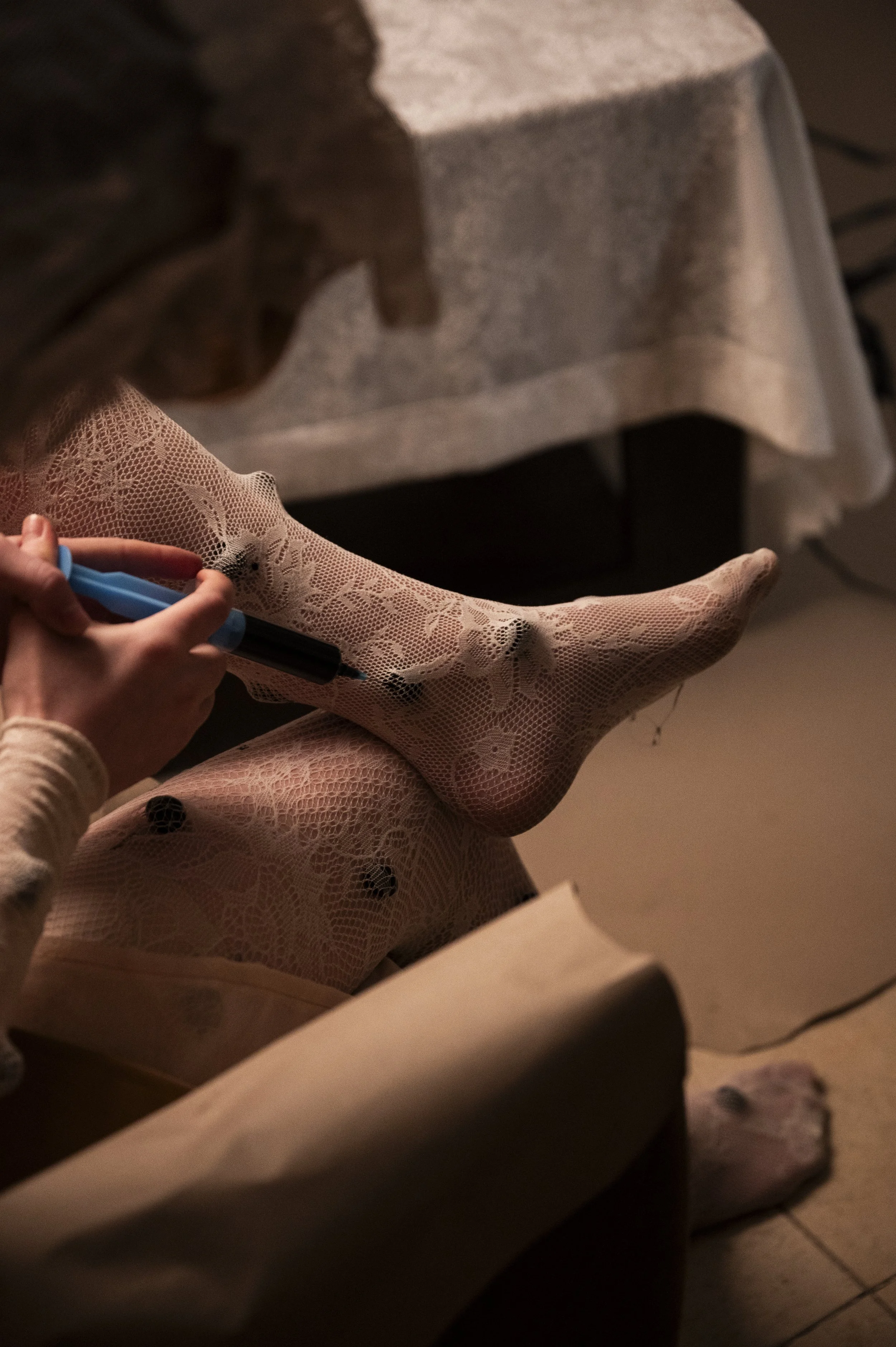 Person sitting with crossed legs wearing lace tights with black dots, using a blue and black marker, in a dimly lit room.